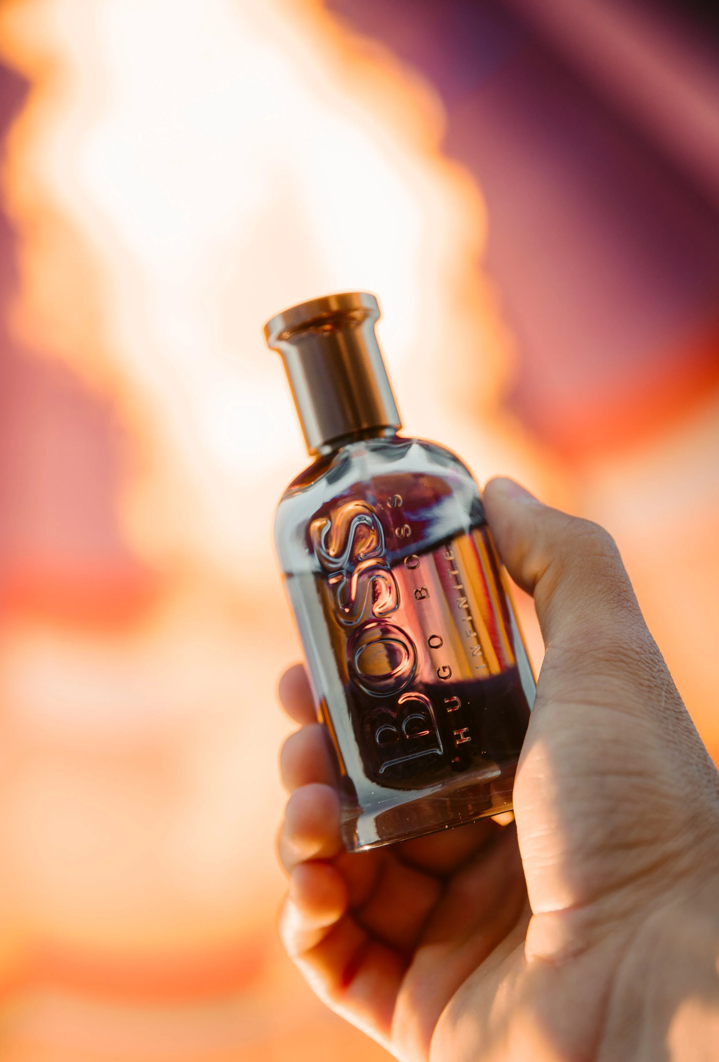 A hand holding a small glass bottle with a metallic cap, with a blurred fire in the background.