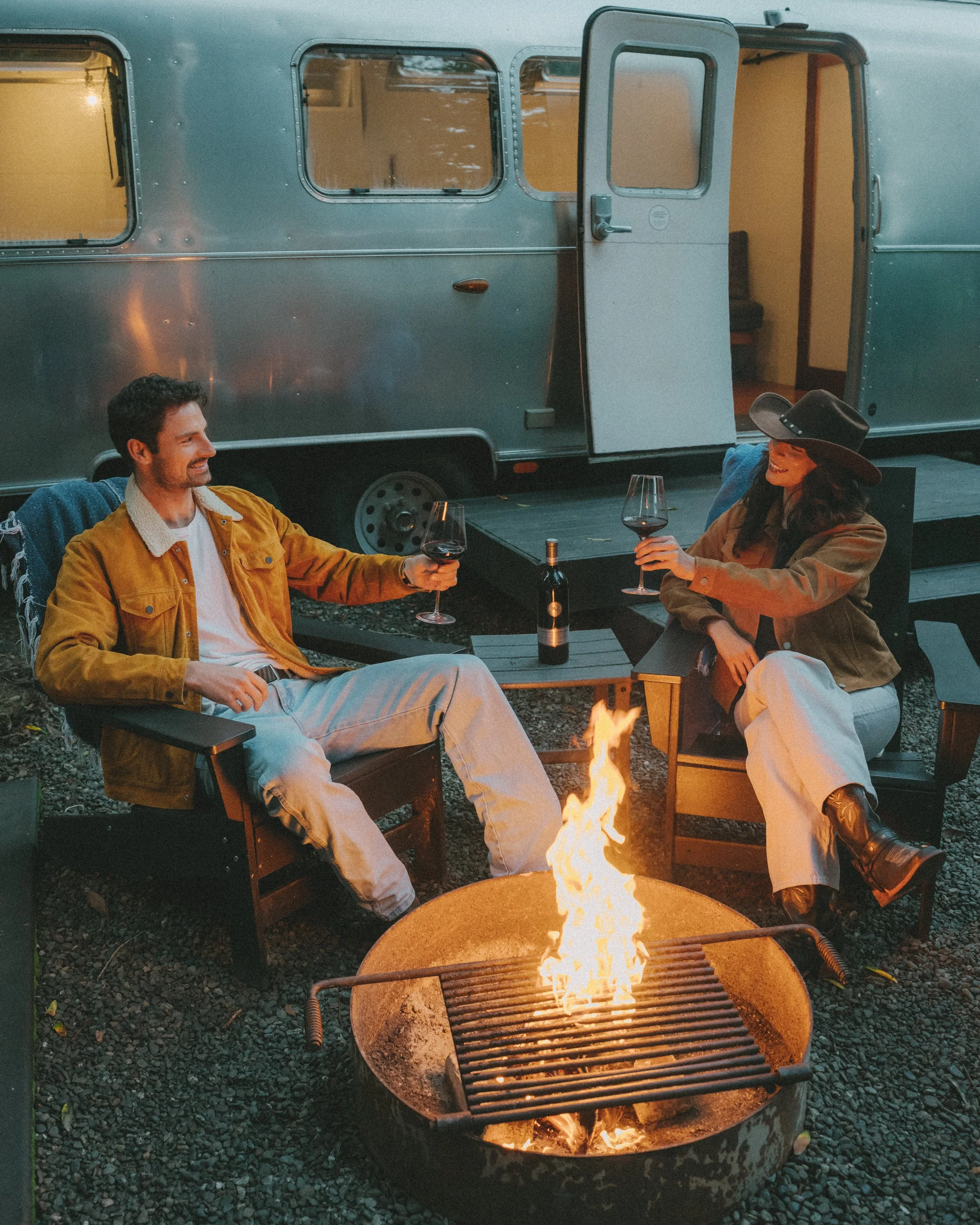 Two people sitting around a campfire outside at night, holding glasses of red wine and smiling. A man in a yellow jacket and a woman wearing a wide-brimmed hat and tan jacket. There is a vintage silver camper trailer in the background with its door o