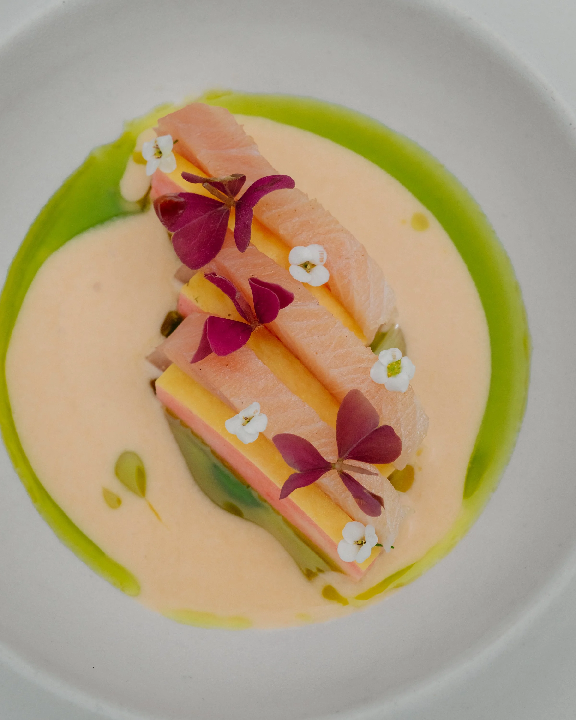 A beautifully decorated dessert with pink and yellow layers, garnished with white and purple edible flowers, placed on a white plate with green accents.
