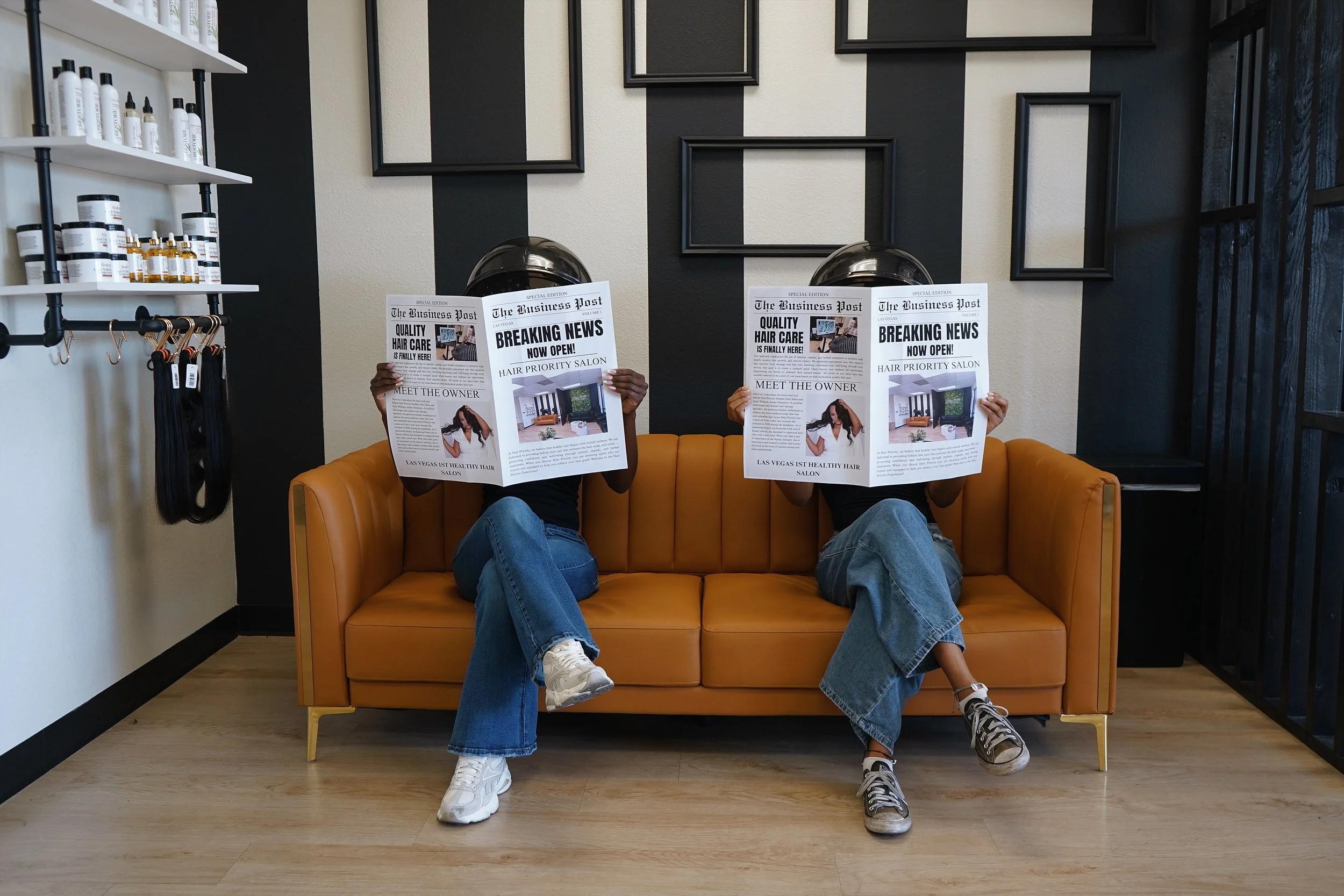 Two people sitting on a yellow sofa in a salon, reading newspapers with only their hands and legs visible. The salon has black and white walls, empty black frames, and shelves with hair products. The floor is wood, and there are hair extensions hanging on the left.