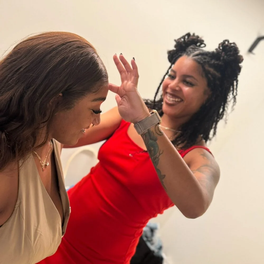 Two women smiling and playfully interacting, one wearing a beige top and the other in a red dress, with the woman in red giving a high-five or playful gesture.