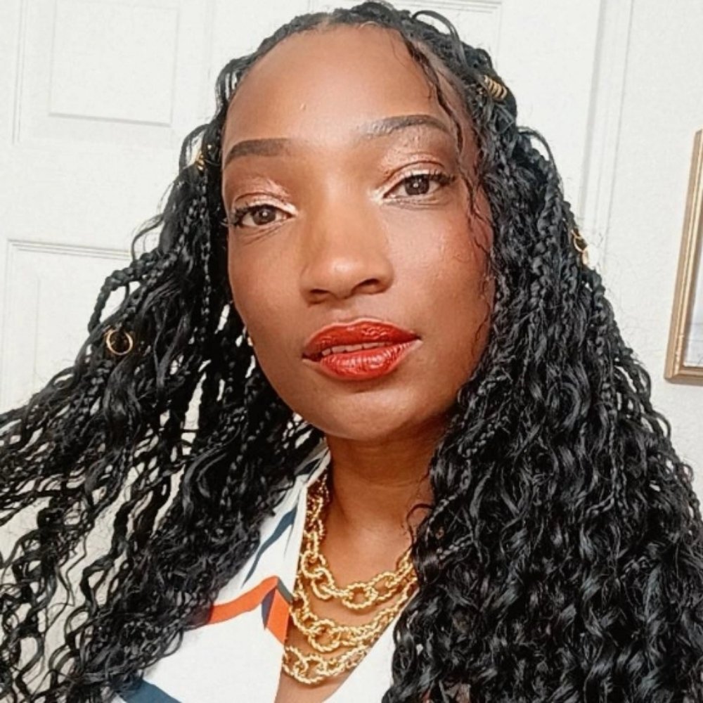 Close-up of an African American woman with curly black hair, wearing gold jewelry, red lipstick, silver eyeshadow, and a white top with orange and navy accents, standing indoors in front of a white door and wall.