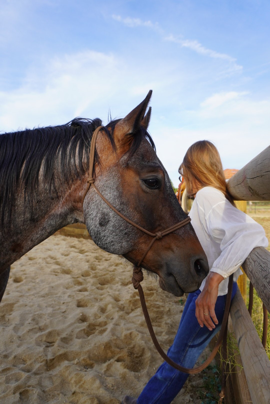 A woman with red hair leaning against a wooden post while a gray and brown horse stands next to her on a sandy horse riding area, with a blue sky and some clouds overhead.