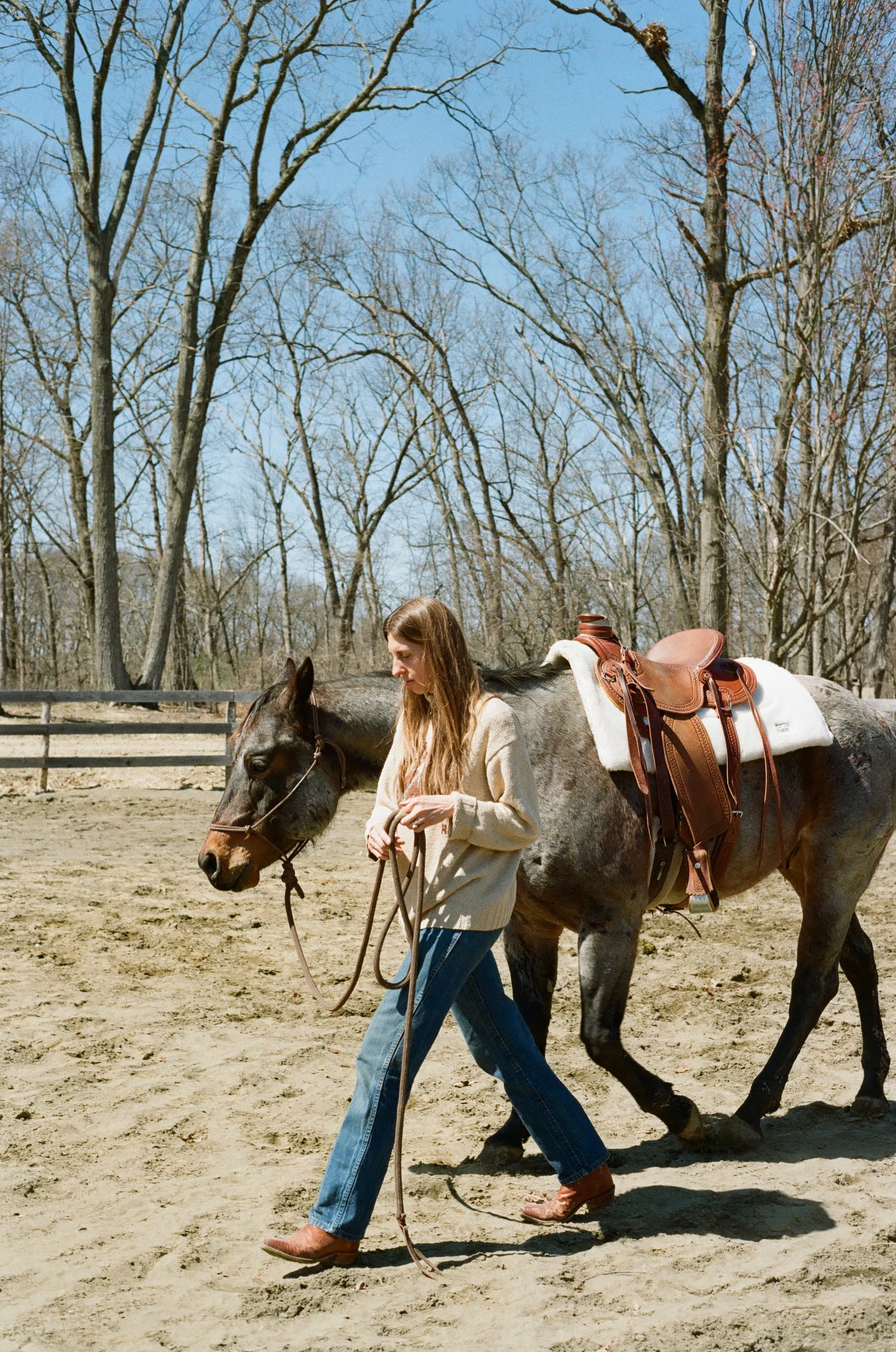 A woman walking a horse on a dirt arena outdoors with leafless trees and a blue sky in the background.
