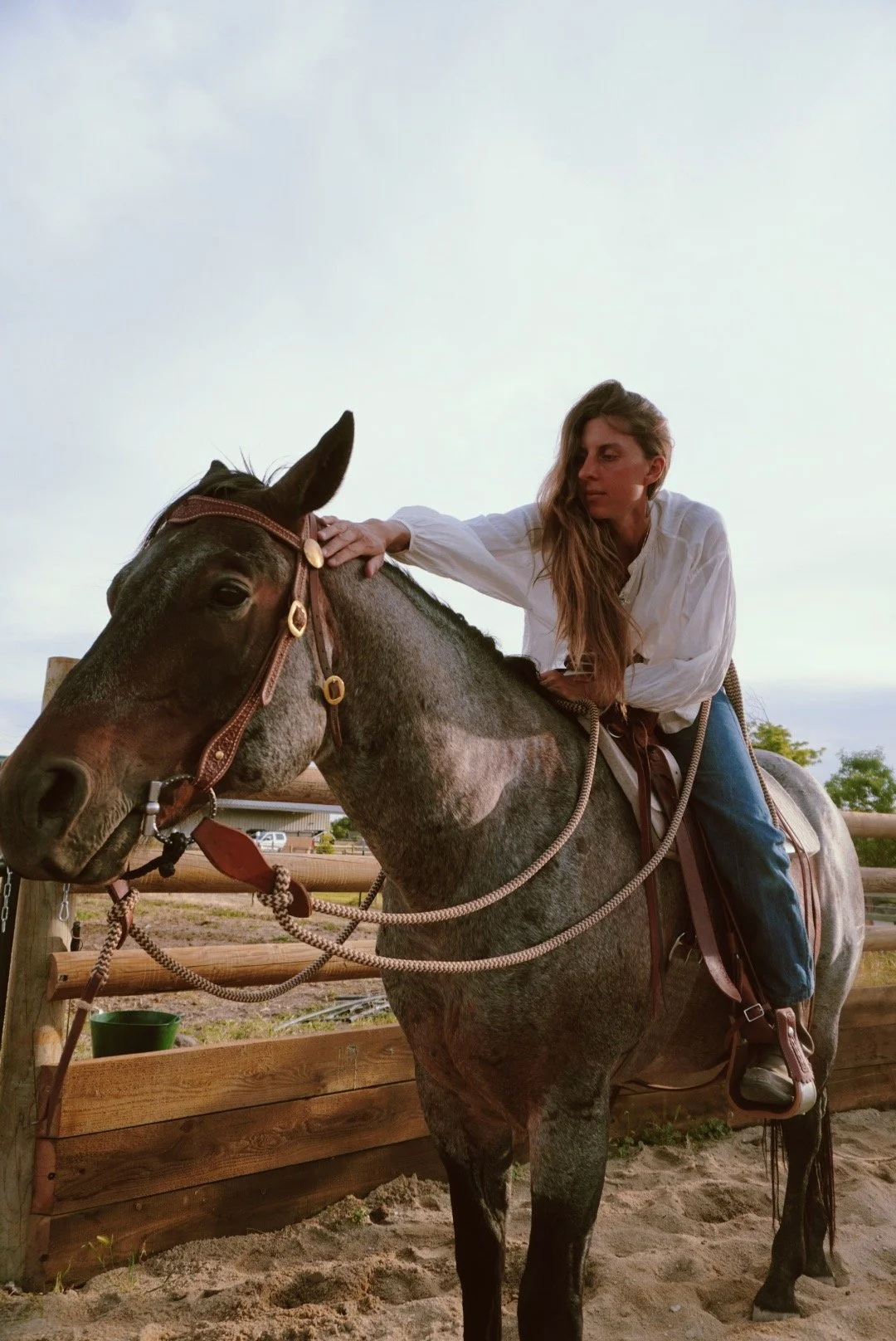 A woman with long hair, wearing a white shirt and blue jeans, riding a gray and brown speckled horse with a brown bridle and saddle in an outdoor riding arena.