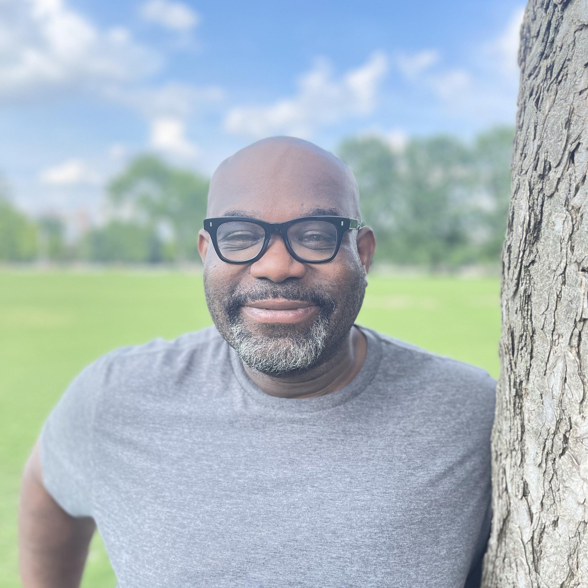A smiling man with glasses partially hidden behind a tree at a park on a sunny day.