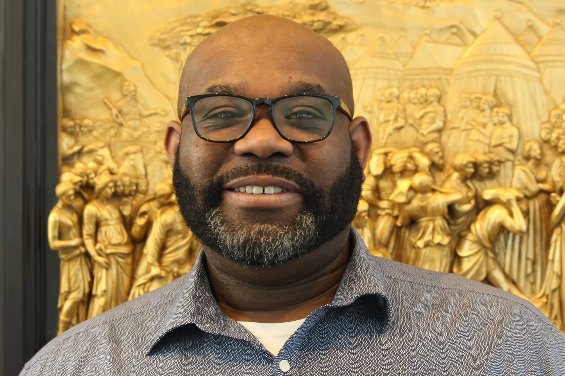 A portrait of a smiling man with glasses and a beard, standing in front of a detailed gold-colored carved wooden artwork depicting a historical scene.