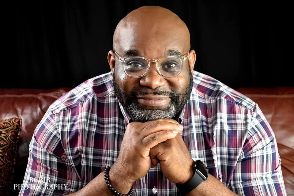 A bald man with a beard and glasses, wearing a plaid shirt, sitting on a brown leather couch with decorative pillows, holding his chin with his hands, and smiling at the camera.