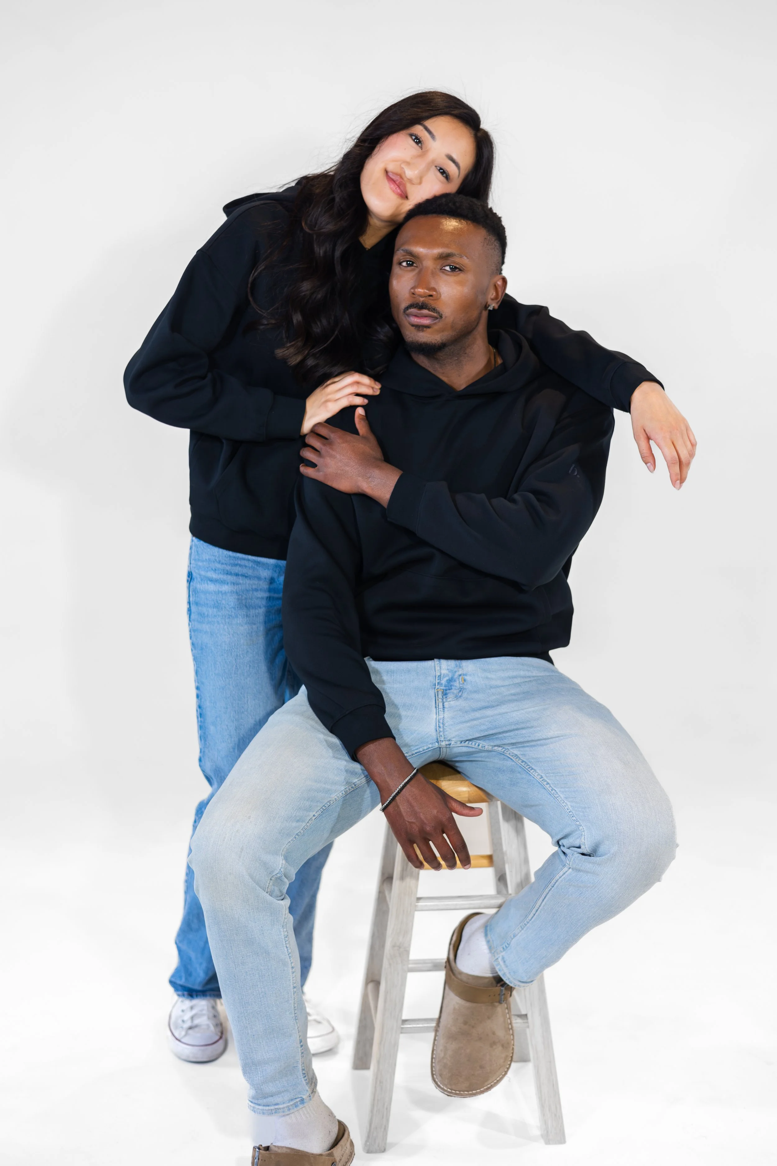 A woman with long dark hair and light skin leaning on a man with short dark hair and darker skin, who is sitting on a stool, against a plain white background. They are wearing casual black hoodies and light blue jeans.