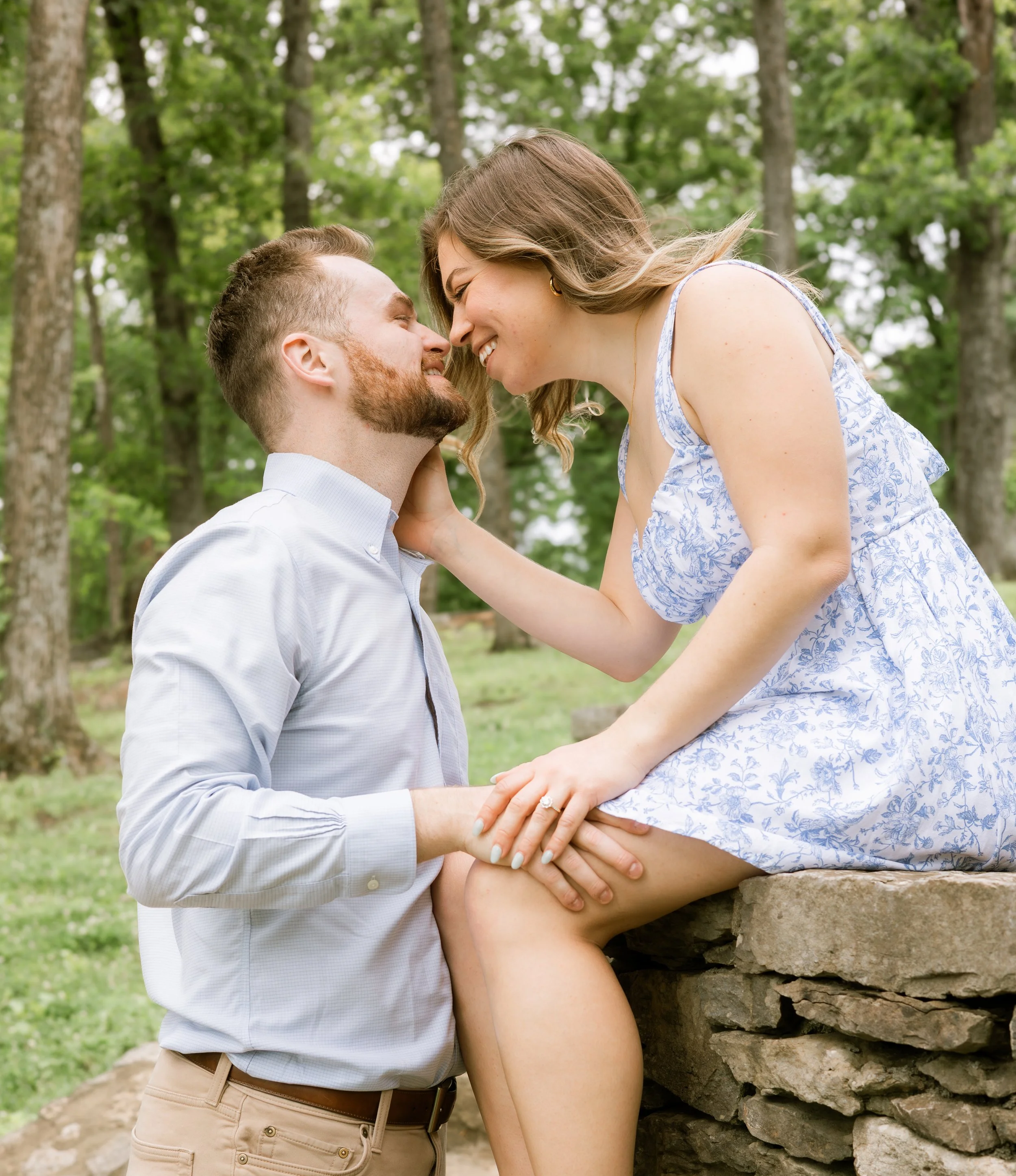 A man with a beard and a woman with long light brown hair are touching foreheads and smiling outdoors in a park with green trees in the background. The woman is sitting on a stone structure, wearing a white and blue floral dress. The man is standing, wearing a white button-up shirt and beige pants.
