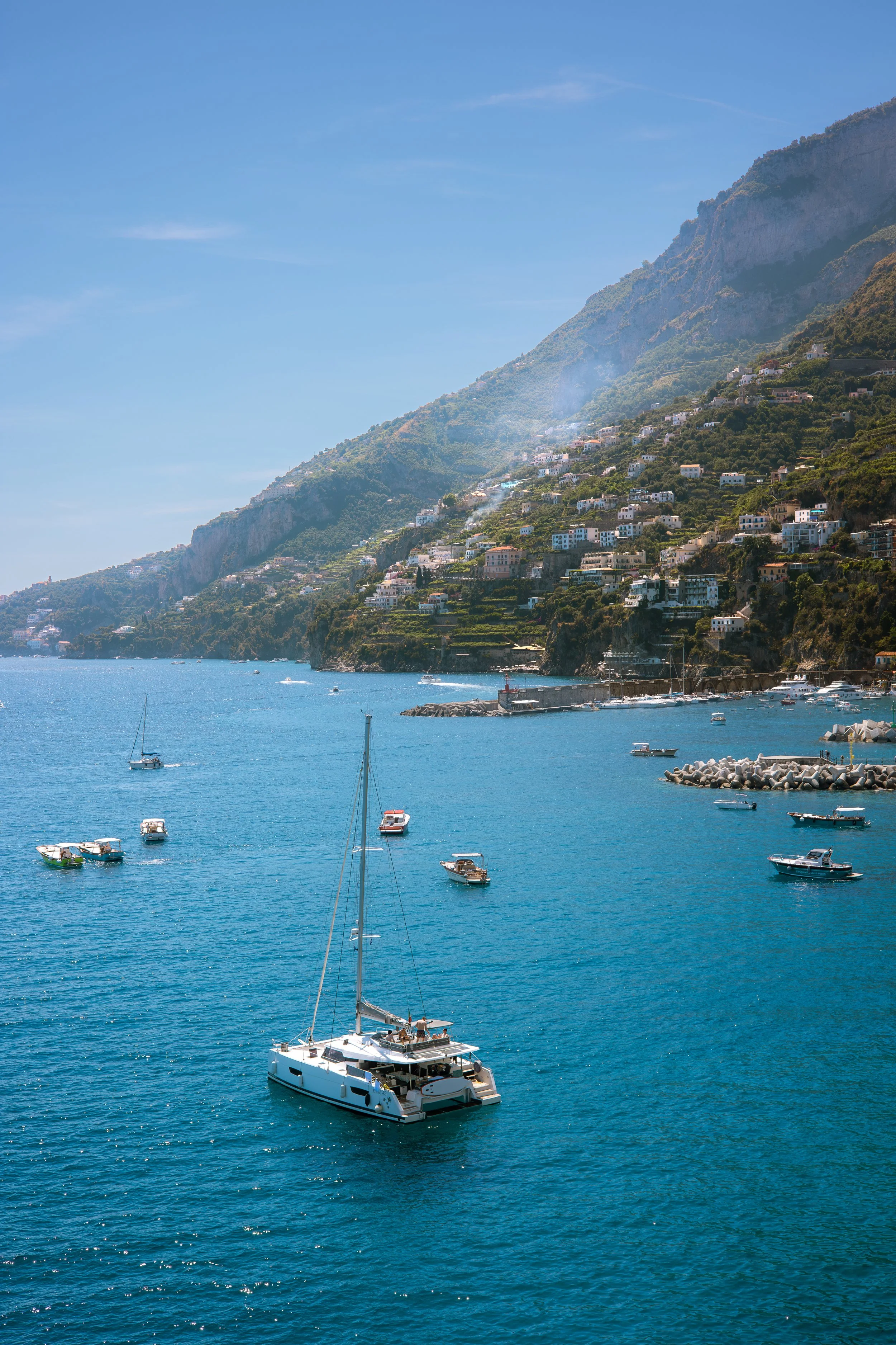 A large white yacht sailing on calm blue water near a coastal hillside with houses and lush greenery under a clear blue sky.