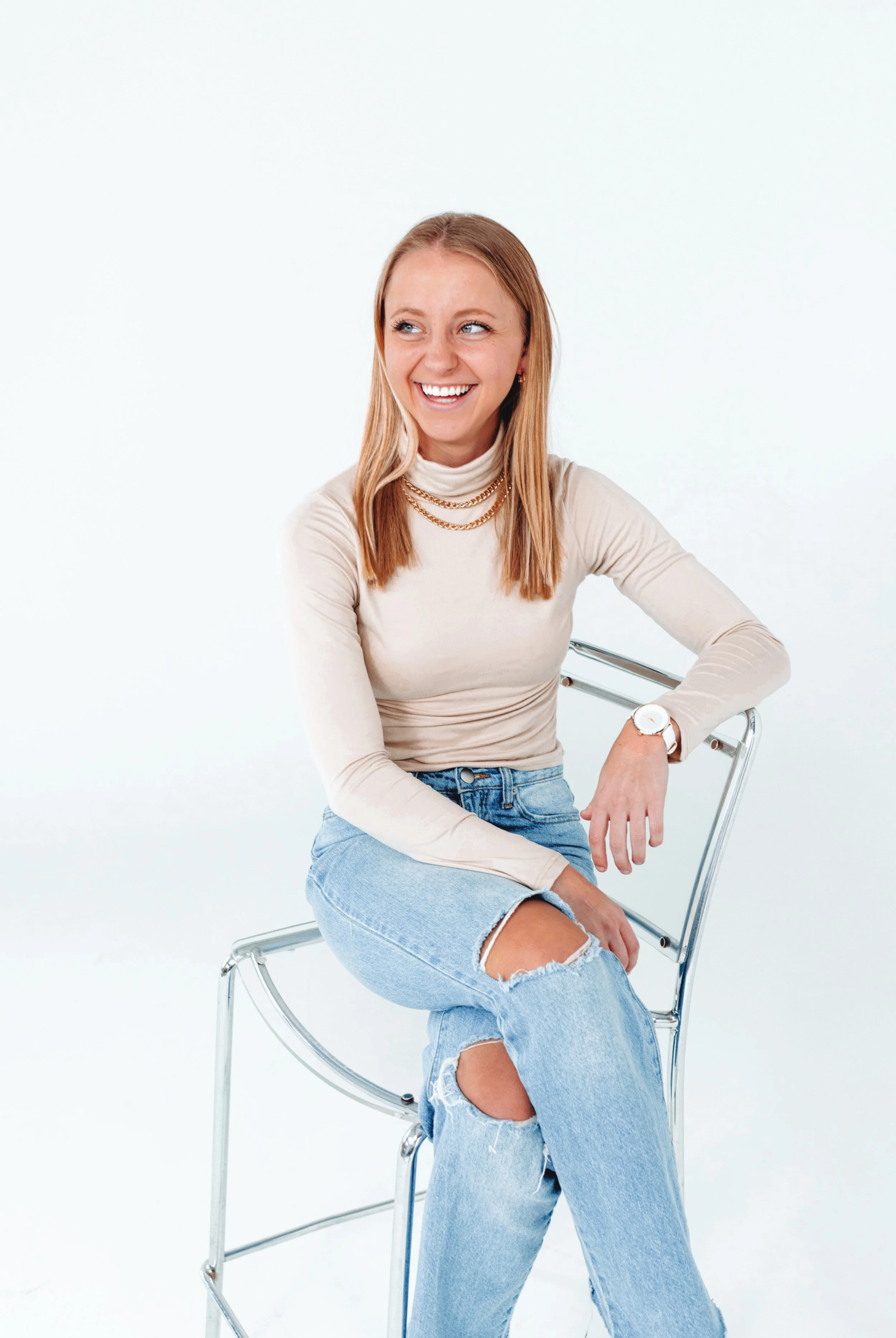 A woman with blonde hair smiling while sitting on a clear acrylic chair against a plain white background, wearing a beige turtleneck sweater, ripped jeans, and layered gold necklaces.
