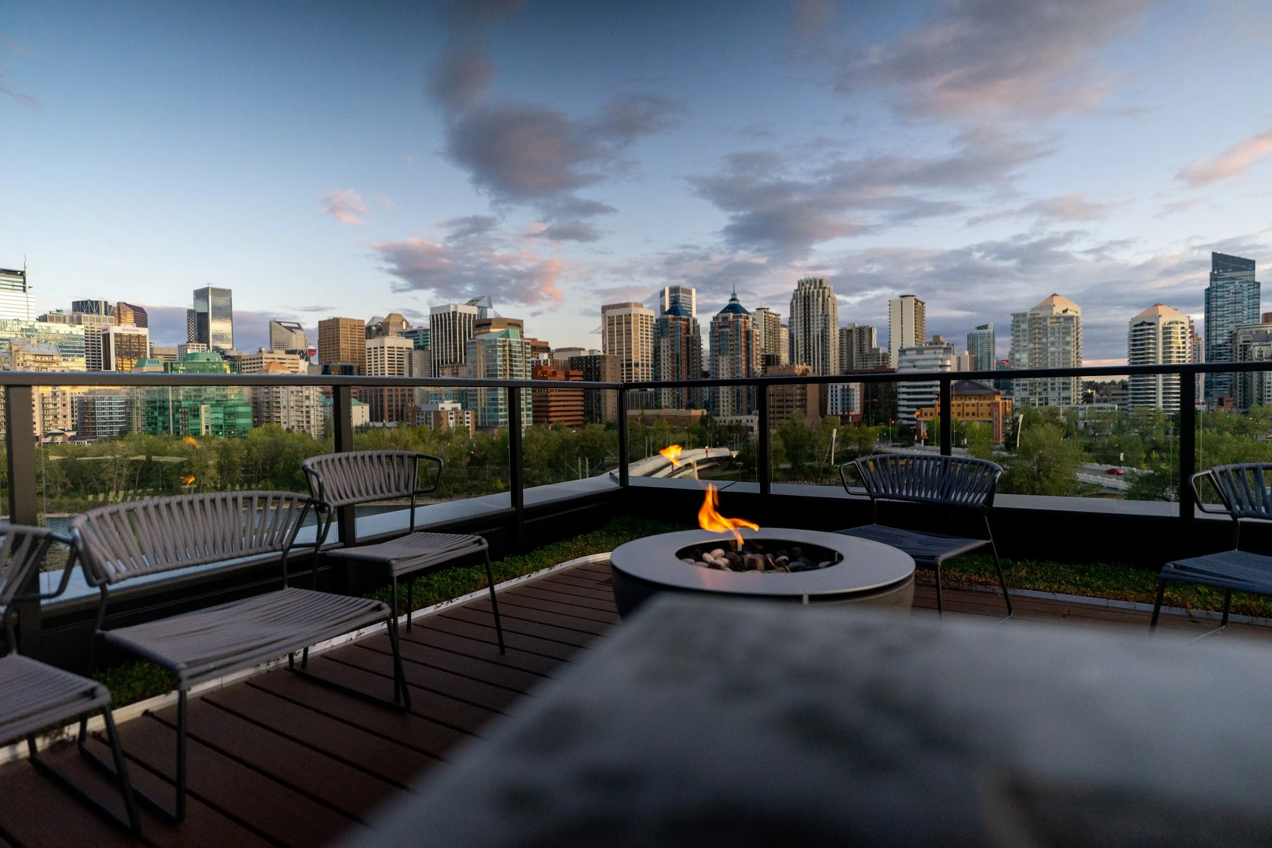 City skyline at sunset viewed from a rooftop patio with outdoor chairs, a fire pit, and a railing. Hive Calgary