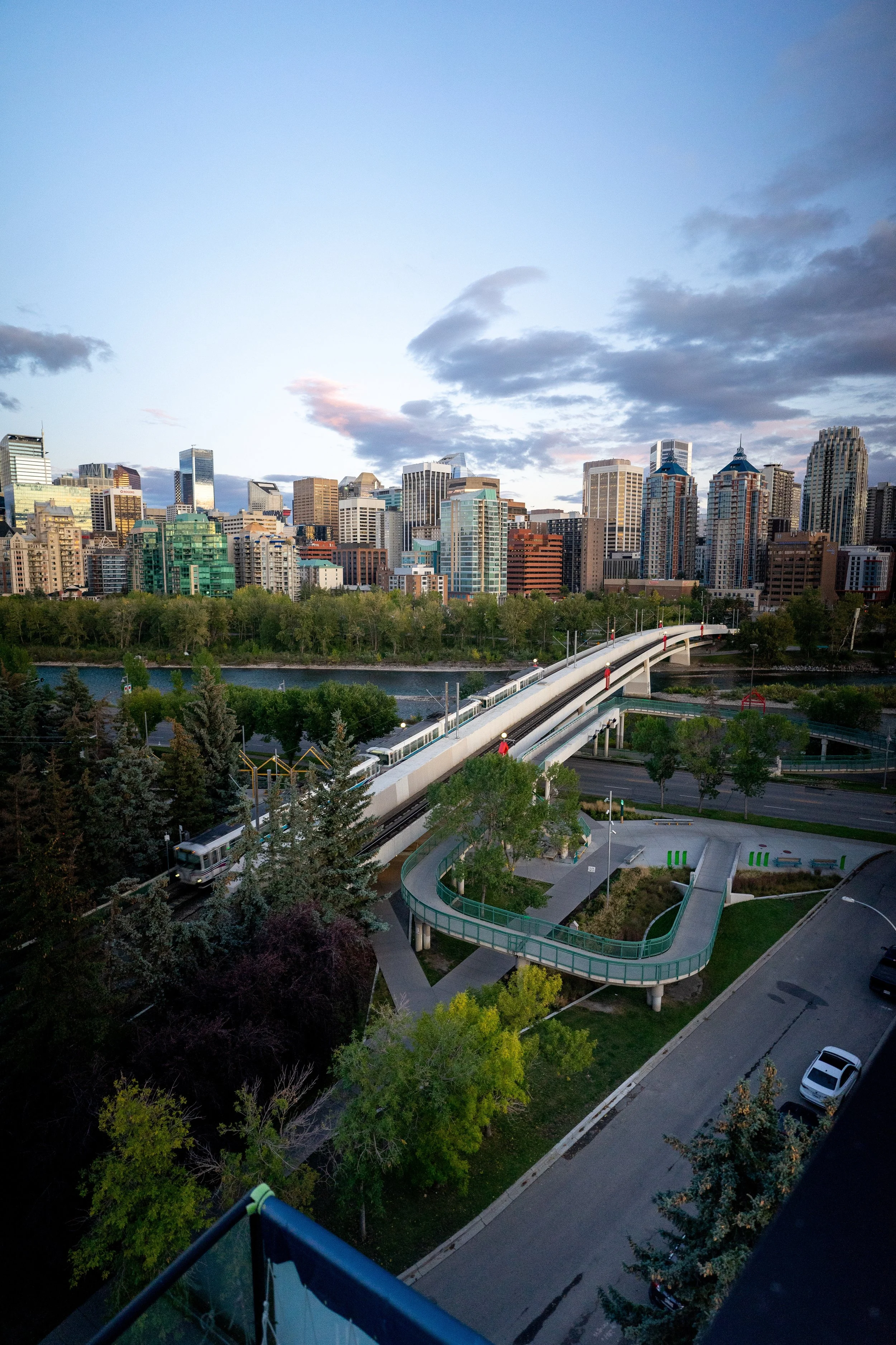 City skyline with numerous tall buildings, a river in the foreground, and a pedestrian bridge over the river. Trees line the riverbank and a street with parked cars is visible in the lower right corner. The sky is partly cloudy.
