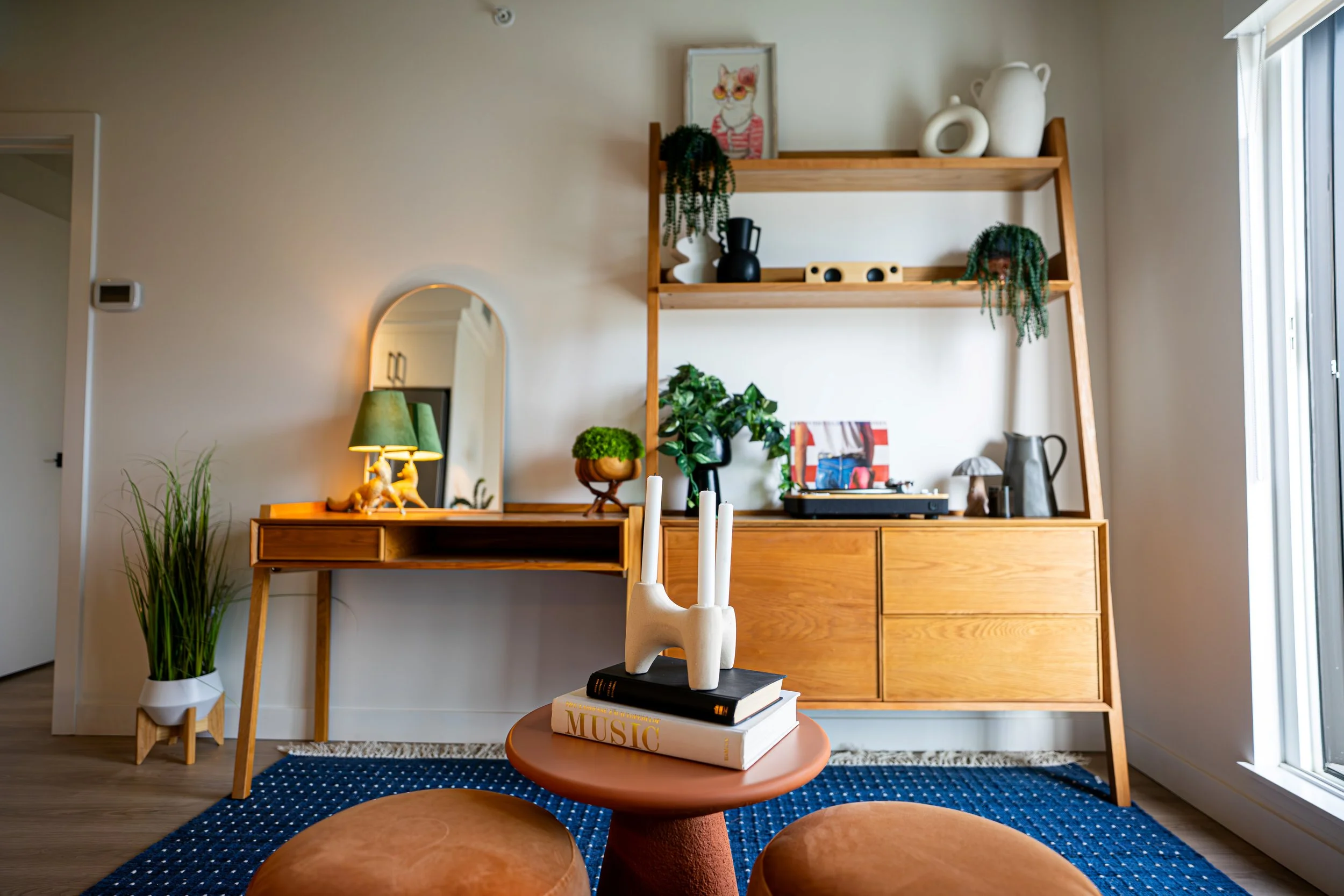 Cozy living room with wooden furniture, decorative vases, plants, artist records, a mirror, green lamp, and colorful artwork, illuminated by natural sunlight.