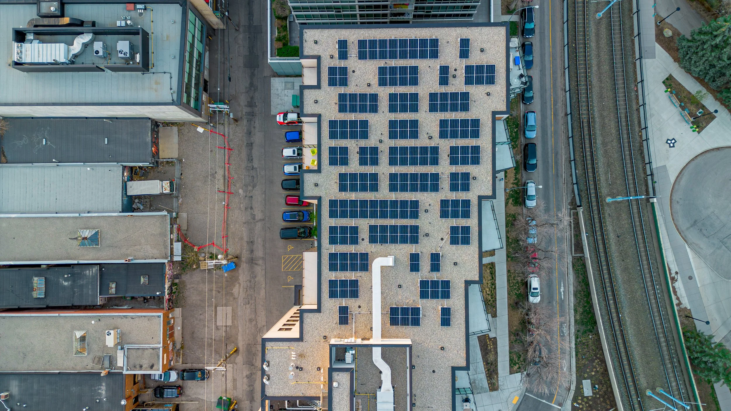 An aerial view of a building with a large rooftop covered with solar panels, surrounded by parking spaces, neighboring buildings, a road, railroad tracks, and a small park area with trees and walkways. Hive Calgary