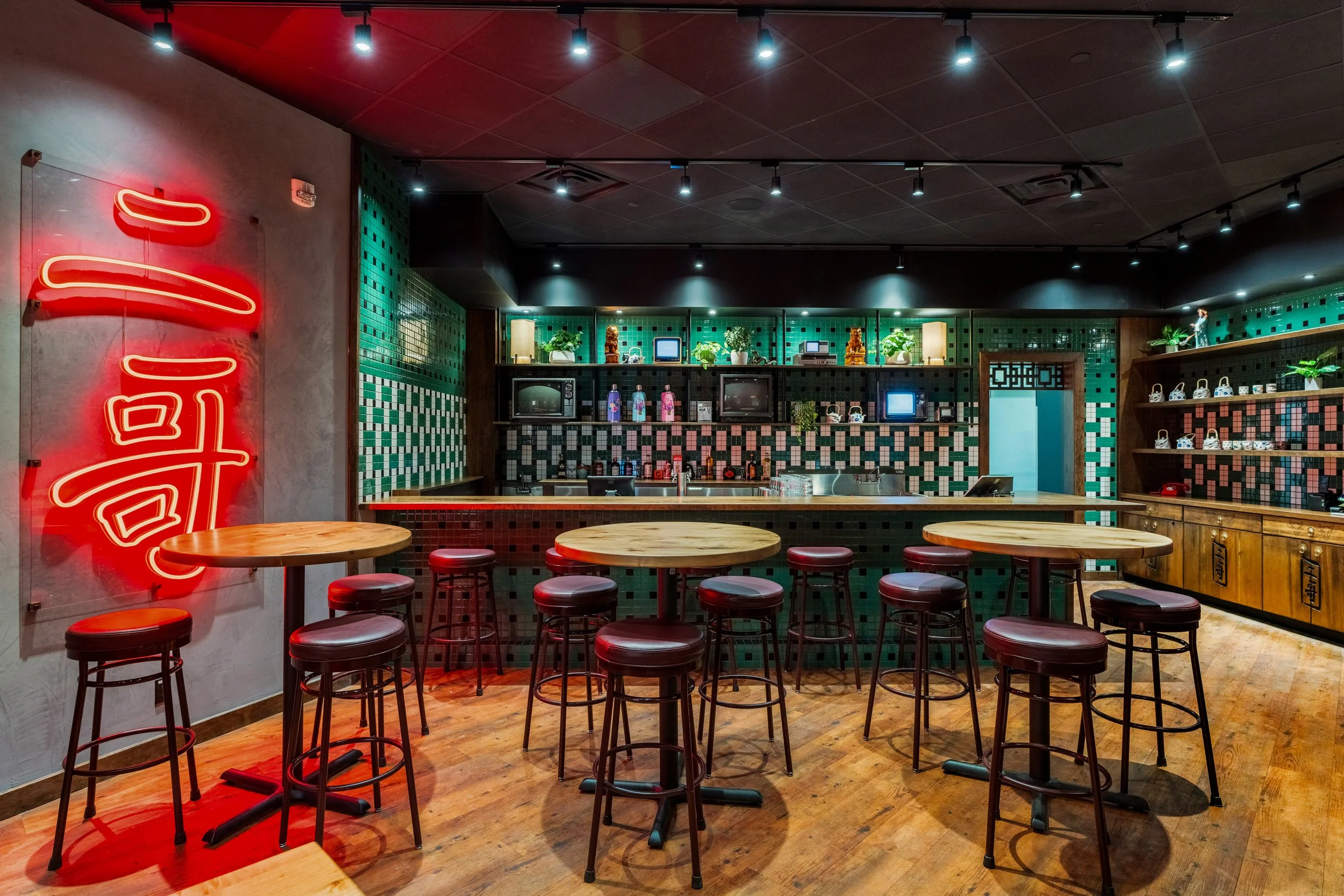 Interior of a restaurant or bar with round wooden tables and red cushioned stools. The back wall has green and black checkered tiles, shelves with plants, vintage TVs, and decorative items. A neon red sign with Asian characters is on the left wall, and the ceiling has black track lighting.