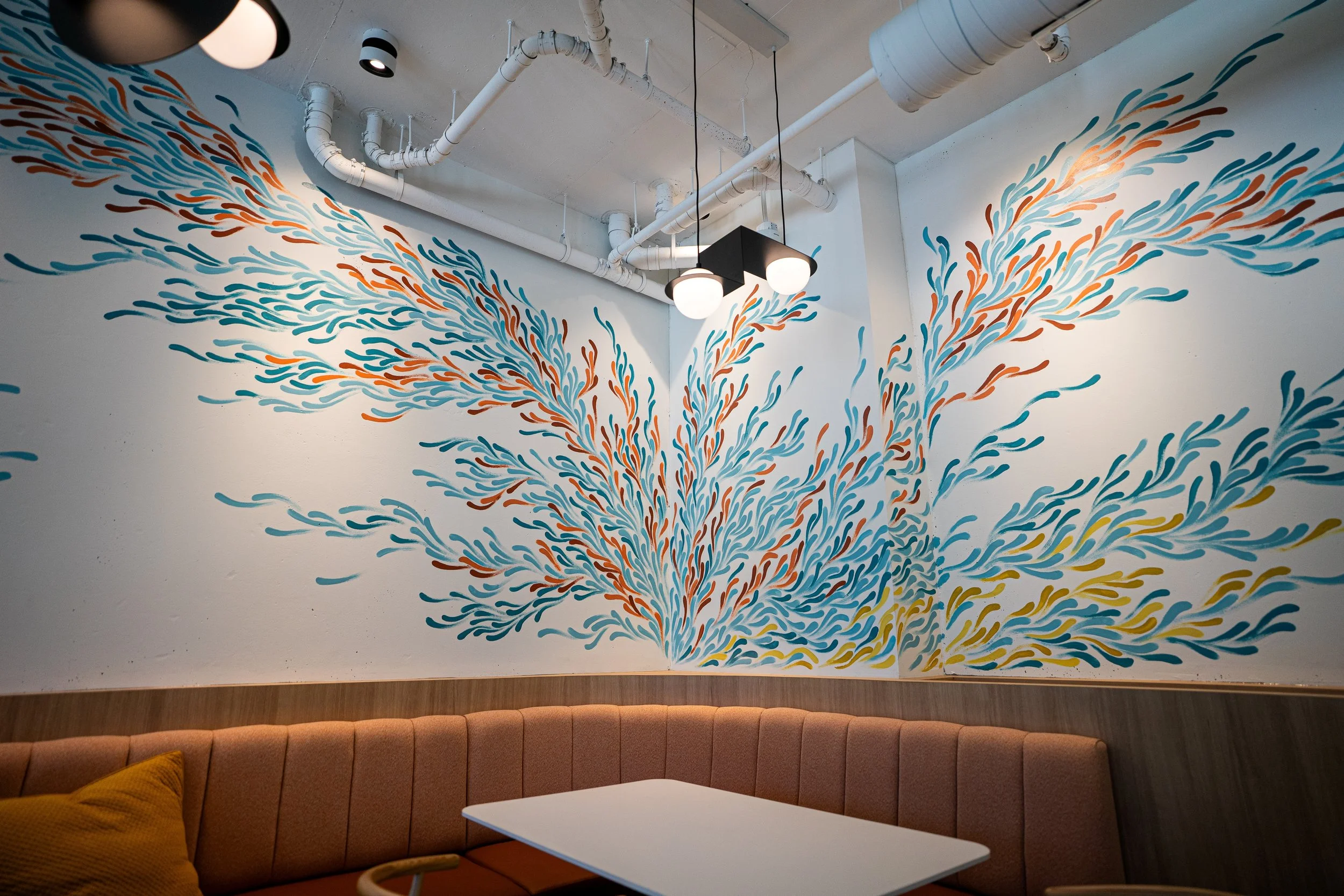 A colorful, abstract wall mural depicting flowing, swirling lines in shades of blue, orange, and yellow, converging towards a corner on the wall behind a round pendant light fixture. The room features an orange upholstered curved bench with mustard-c