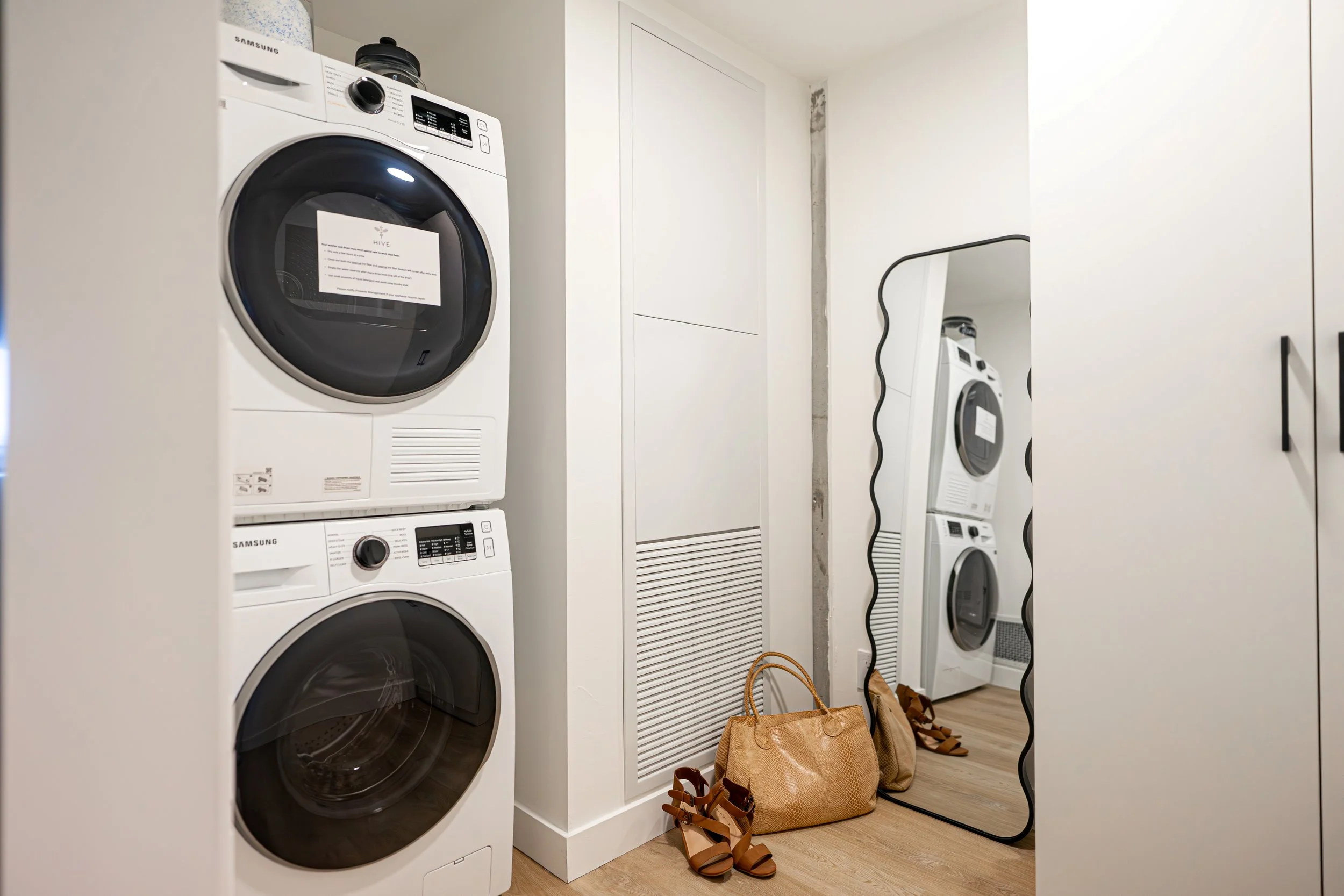 A laundry room with stacked washing machine and dryer, white cabinets, a mirror leaning against the wall, and shopping bags with shoes in front.