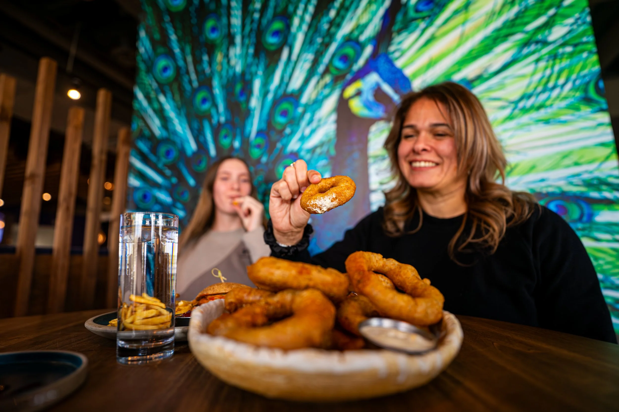 Two women at a restaurant table with a large plate of onion rings, a glass of water, and a side of fries. One woman is smiling and holding an onion ring, the background features a colorful peacock-themed mural.