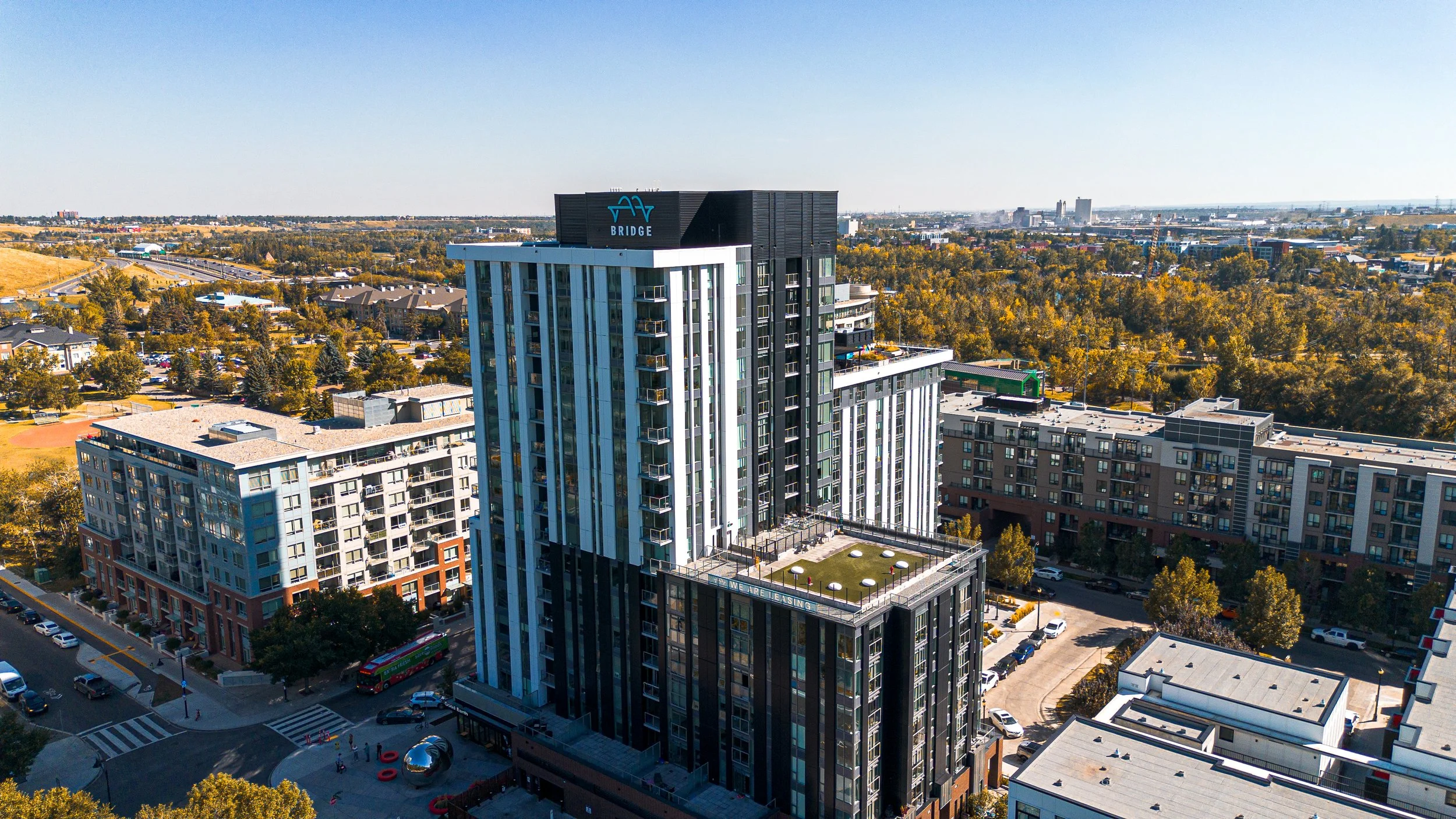 Aerial view of a cityscape featuring a tall modern buildingwith a sign on top that reads 'BRIDGE' and a logo. Surrounding the building are other high-rise residential buildings, roads with cars and buses, a park area, and a distant landscape with trees, hills, and more buildings under a clear blue sky.