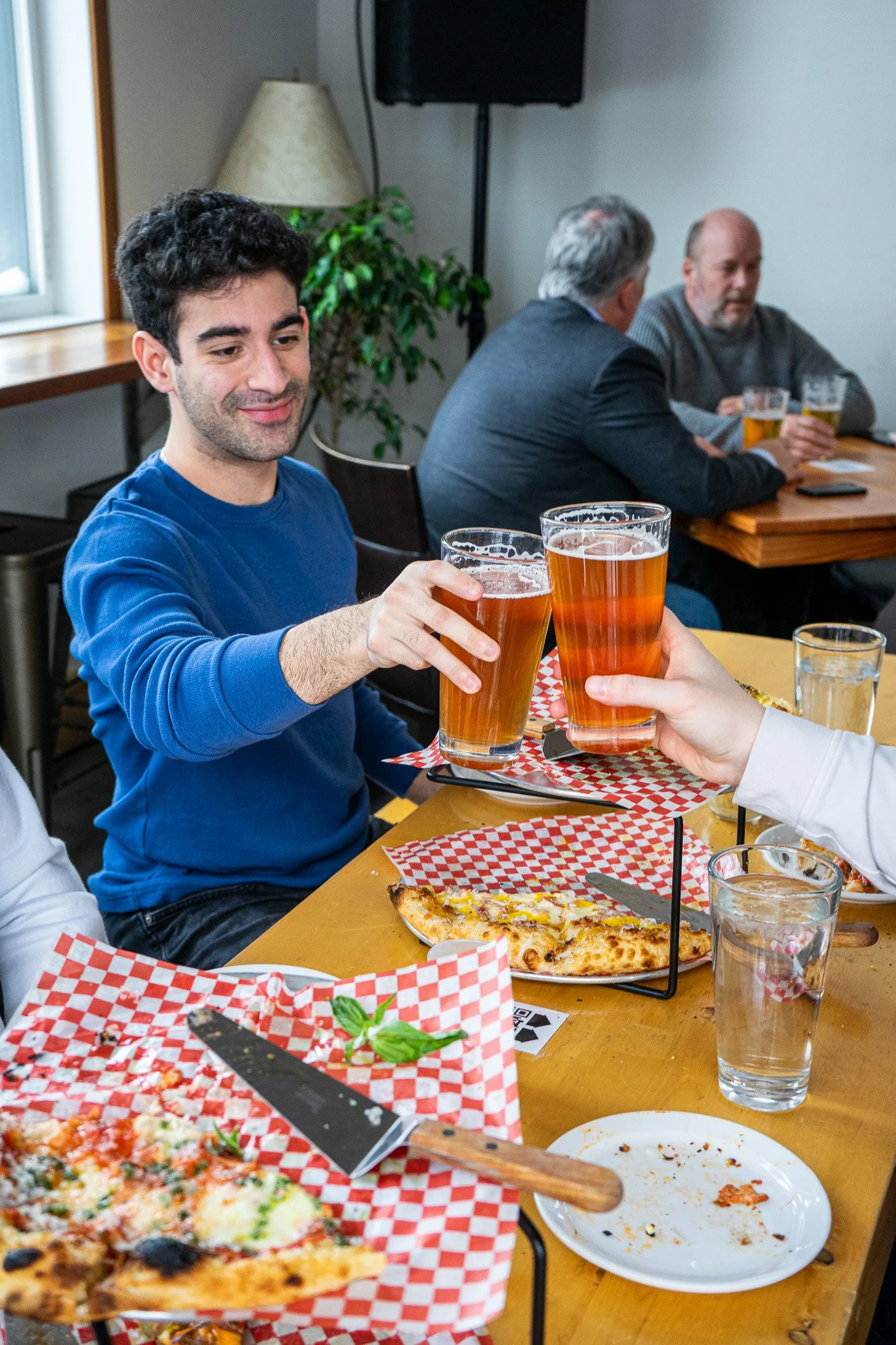 A man in a blue sweater sitting at a table toasting with two glasses of beer with others in the background. There is a pizza on the table and some empty plates. ACME Pizza