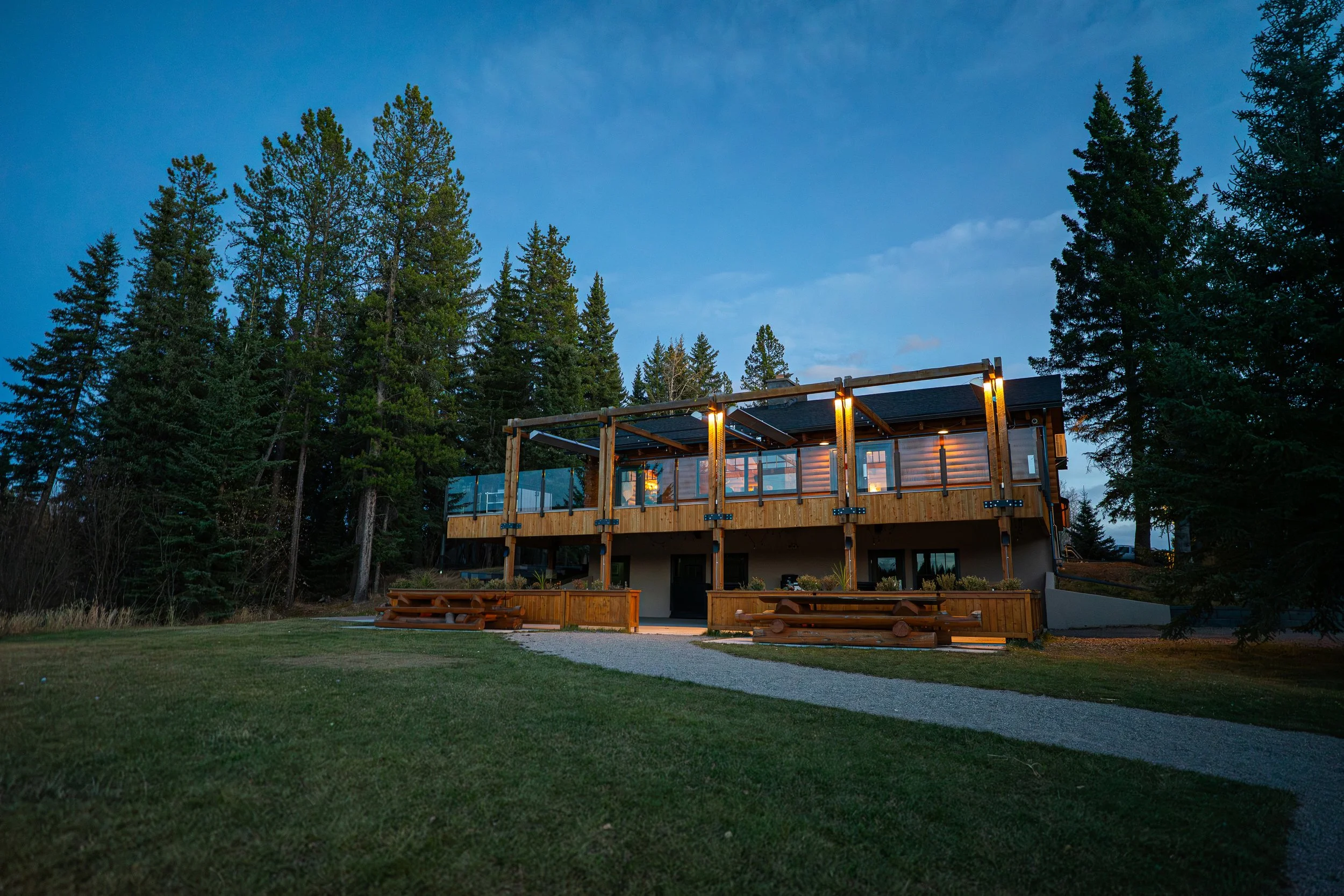 A modern house with wooden accents and large glass windows, situated among tall pine trees under a twilight sky. The house features an elevated deck with wooden railings, outdoor lighting, and picnic tables on the lawn. Schotts Lake