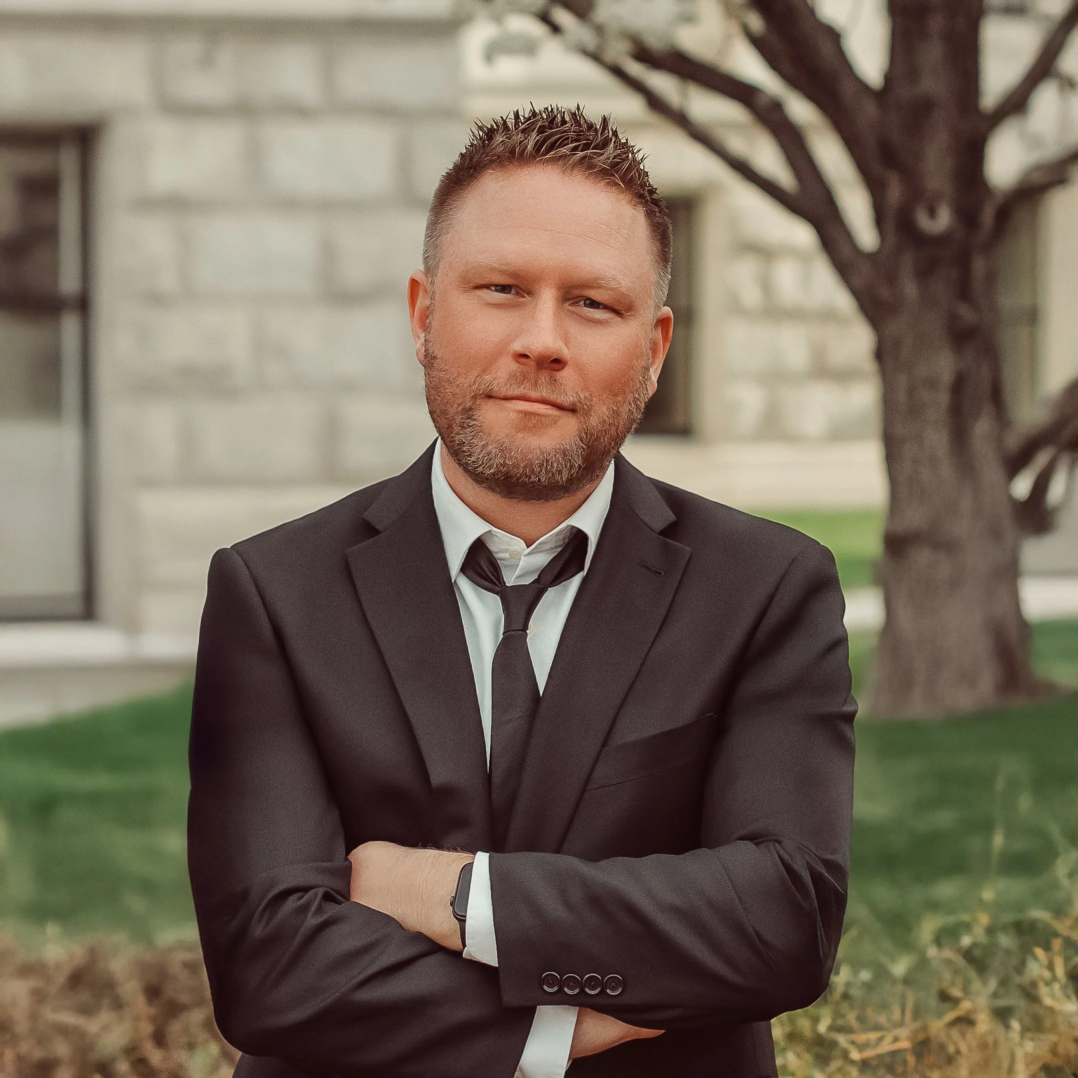 Jim Detjen, a man with short hair and a beard, wearing a black suit, white shirt, and black tie, standing outdoors near a tree and building.