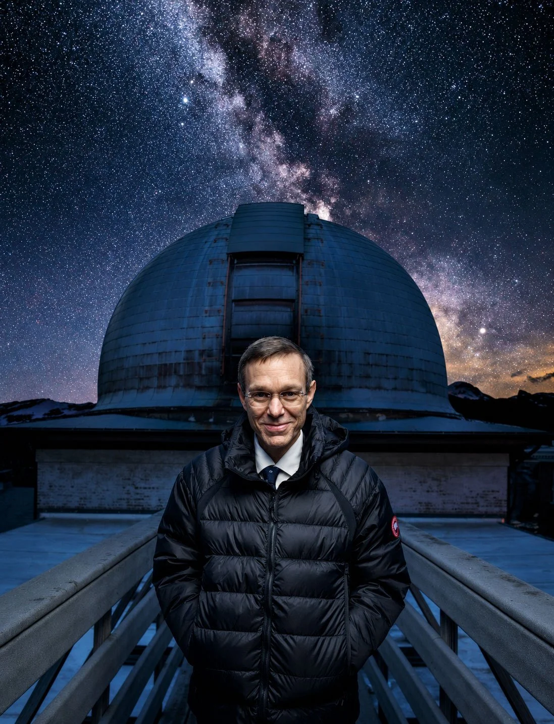 A man wearing glasses and a black jacket stands on a walkway in front of a large observatory with a domed roof. The sky above is filled with stars and the Milky Way galaxy.