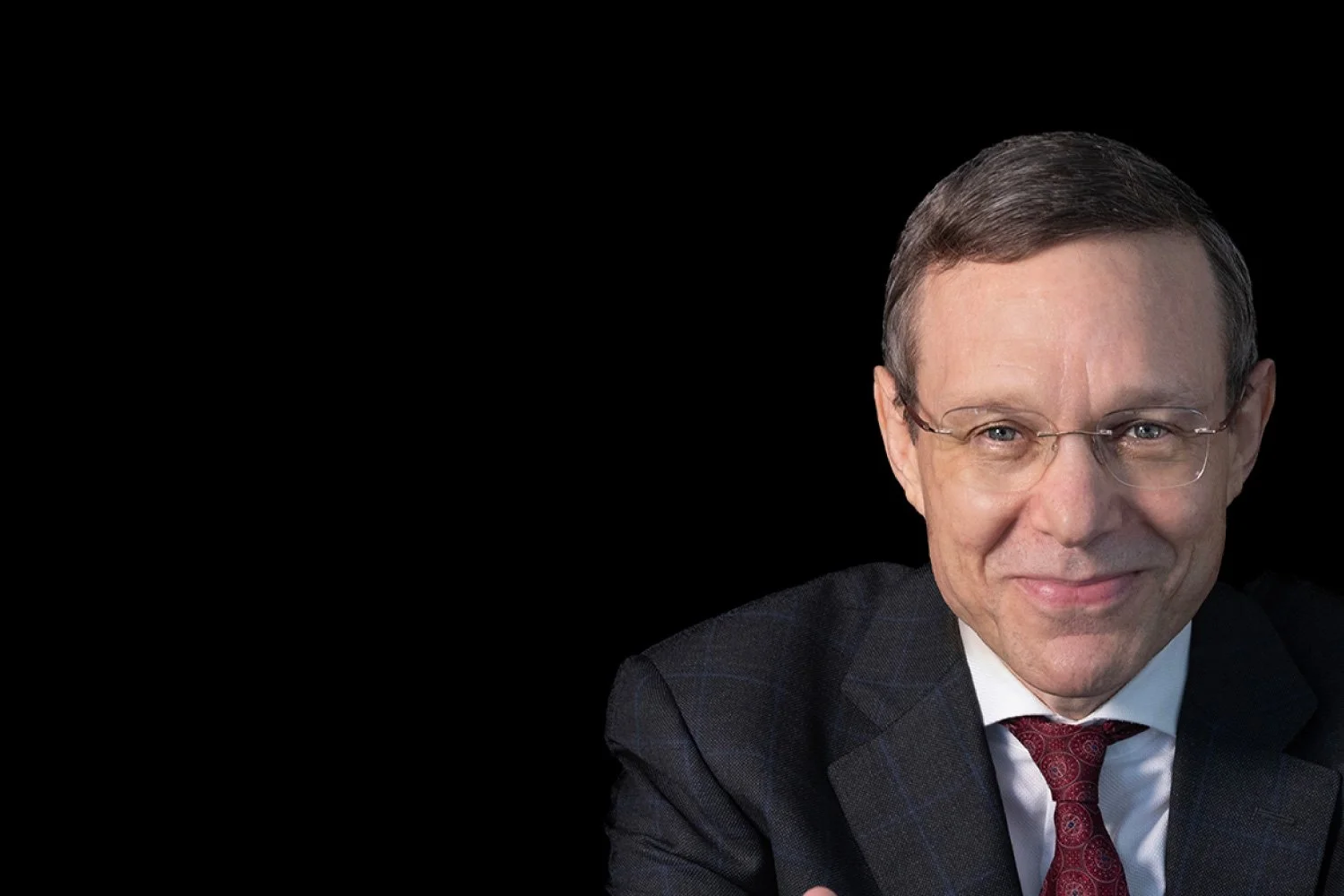 A man in a dark suit, white shirt, and red tie with glasses, smiling at the camera against a plain black background.