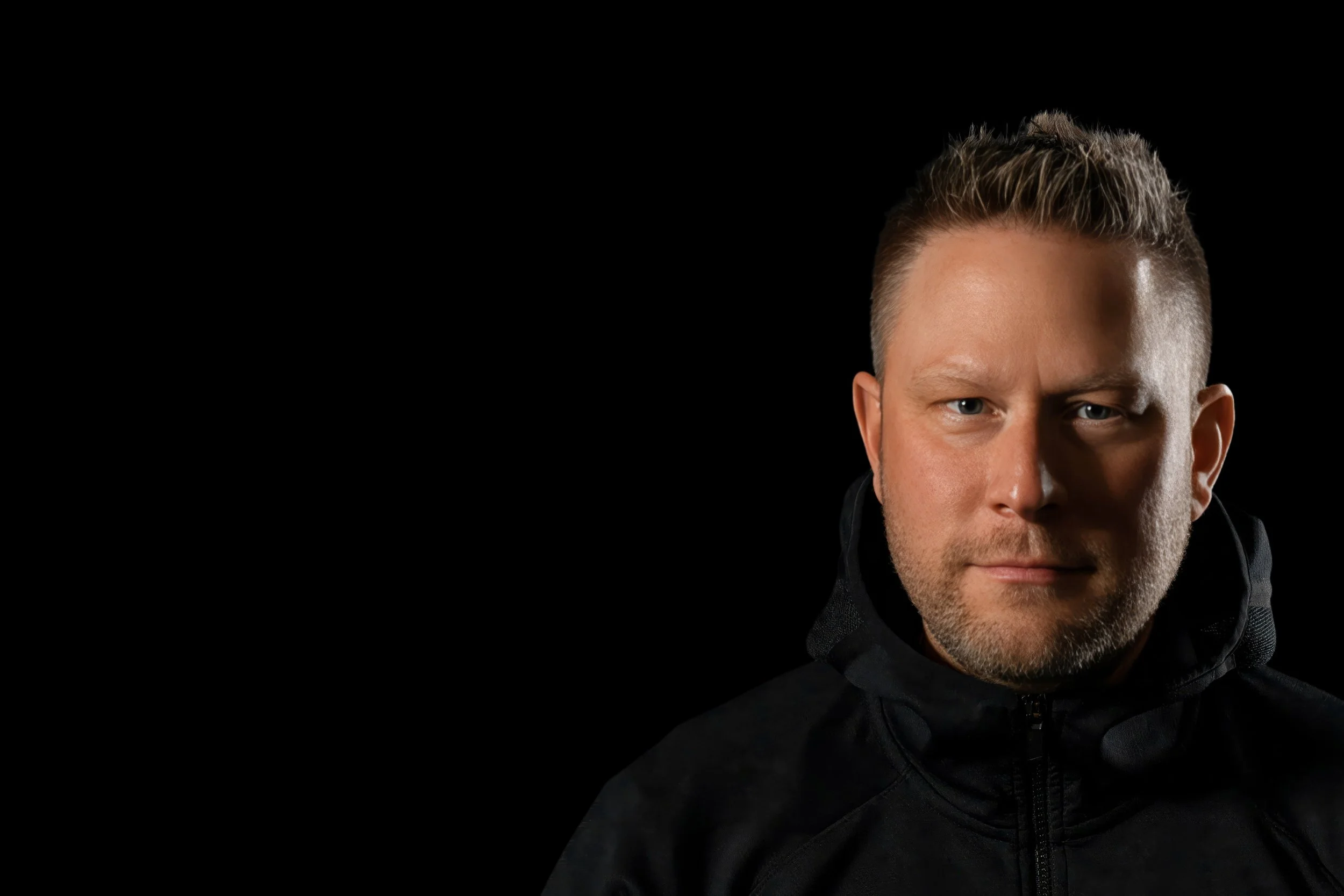 Close-up of a man with short brown hair, blue eyes, and light stubble, wearing a black jacket with a zipper, against a black background.