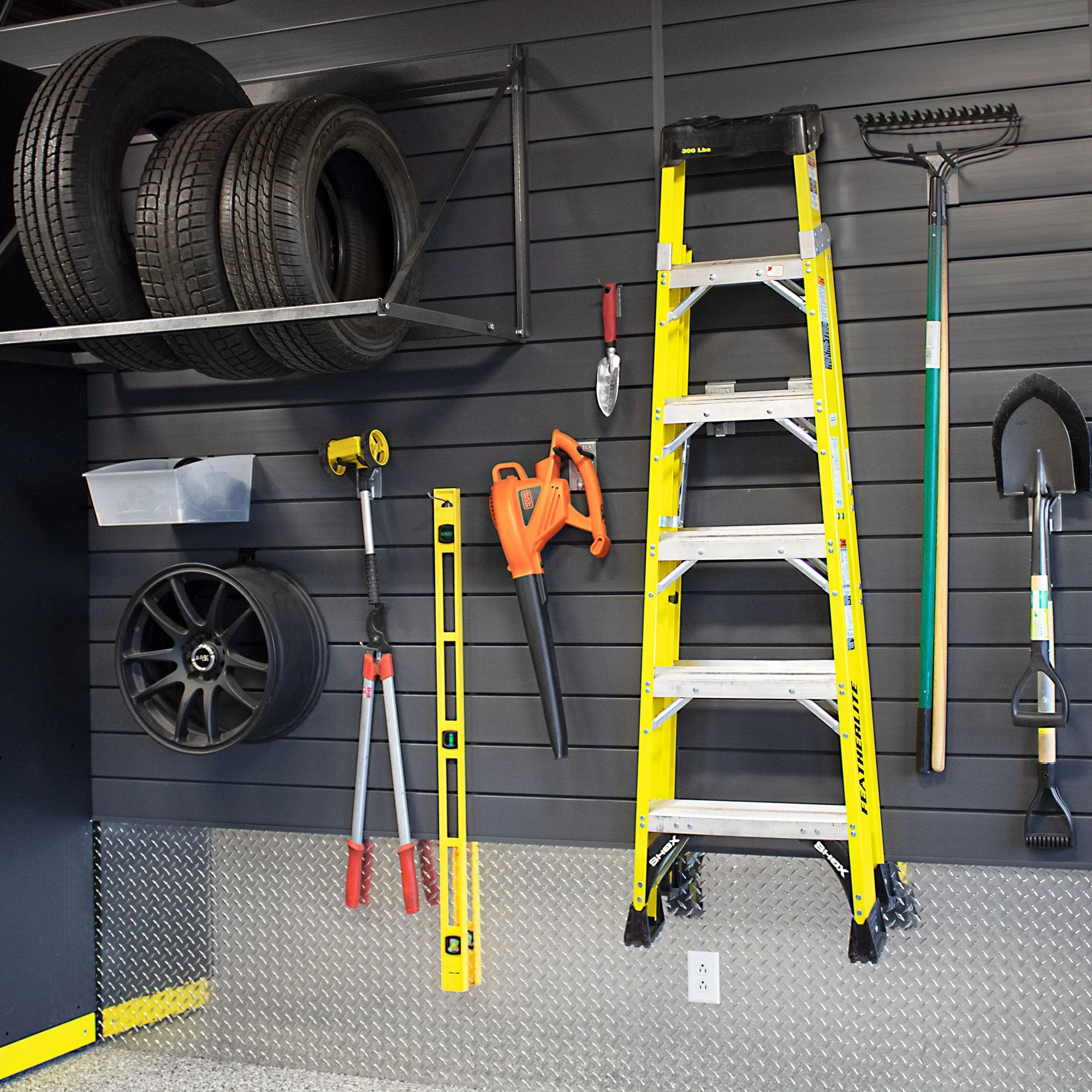 Garage wall with car tires on a metal rack, a yellow step ladder, gardening tools, a hanging shovel, a hedge trimmer, a yellow tape measure, a garden rake, and electrical outlets.