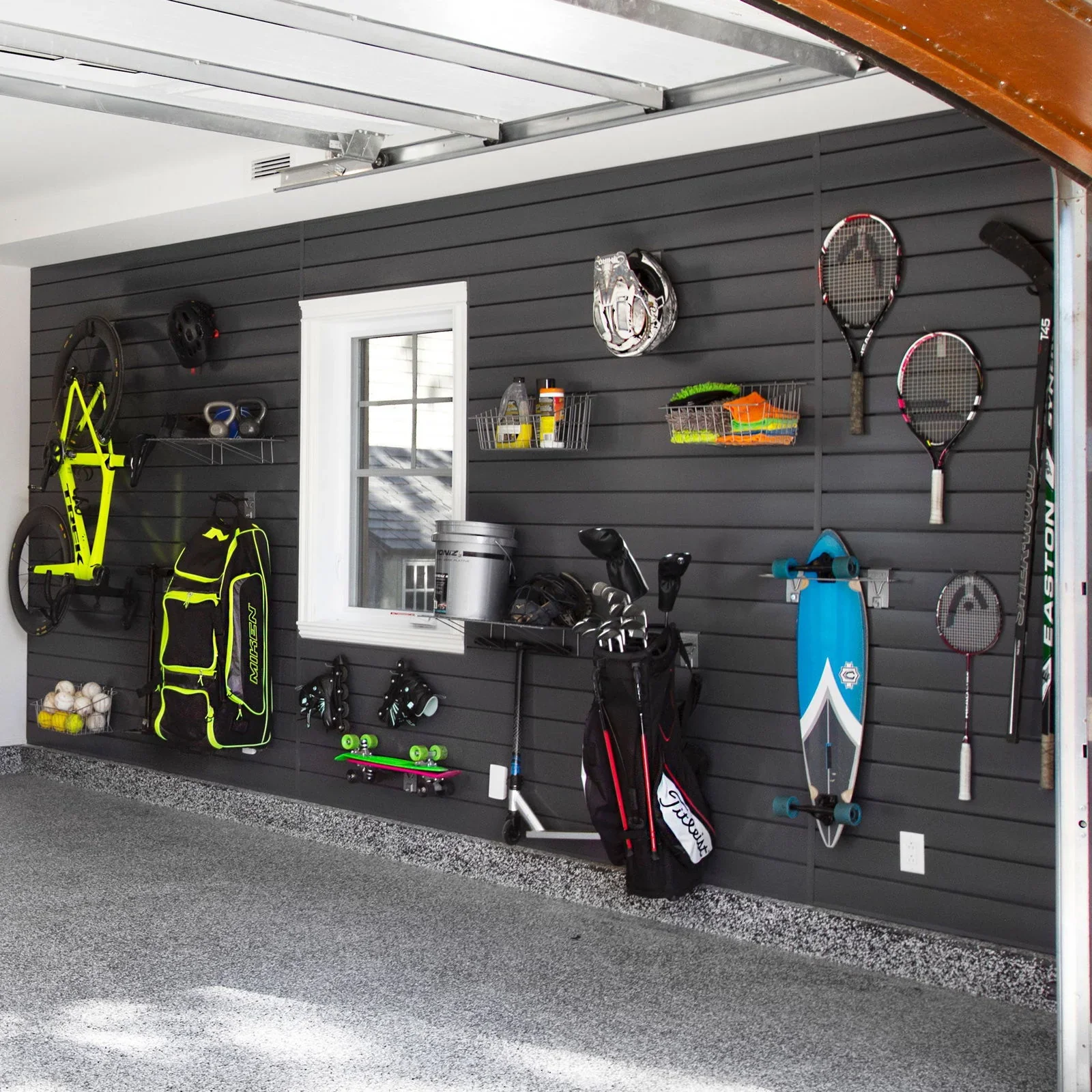 Garage with sports equipment on a black wall, including tennis rackets, helmets, a bike, golf clubs, a skateboard, and a golf bag with clubs, along with tennis balls and a spray bottle on shelves.
