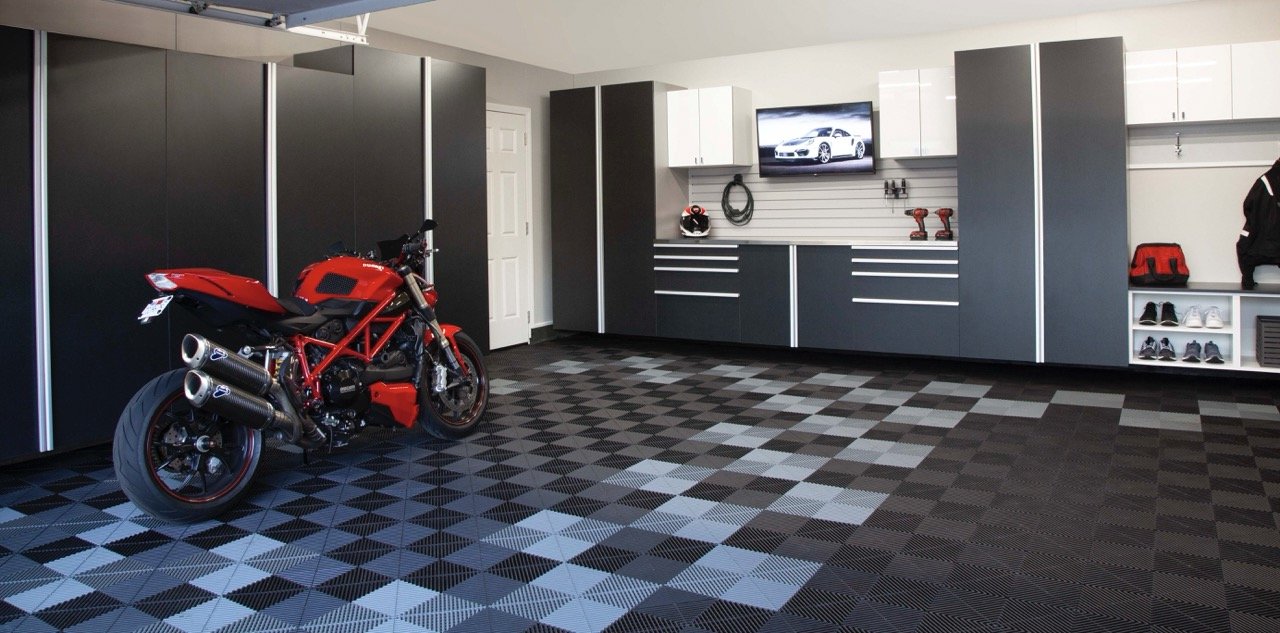 A red motorcycle parked in a garage with black and gray checkered floor tiles, gray cabinets, and a wall-mounted TV displaying a car.