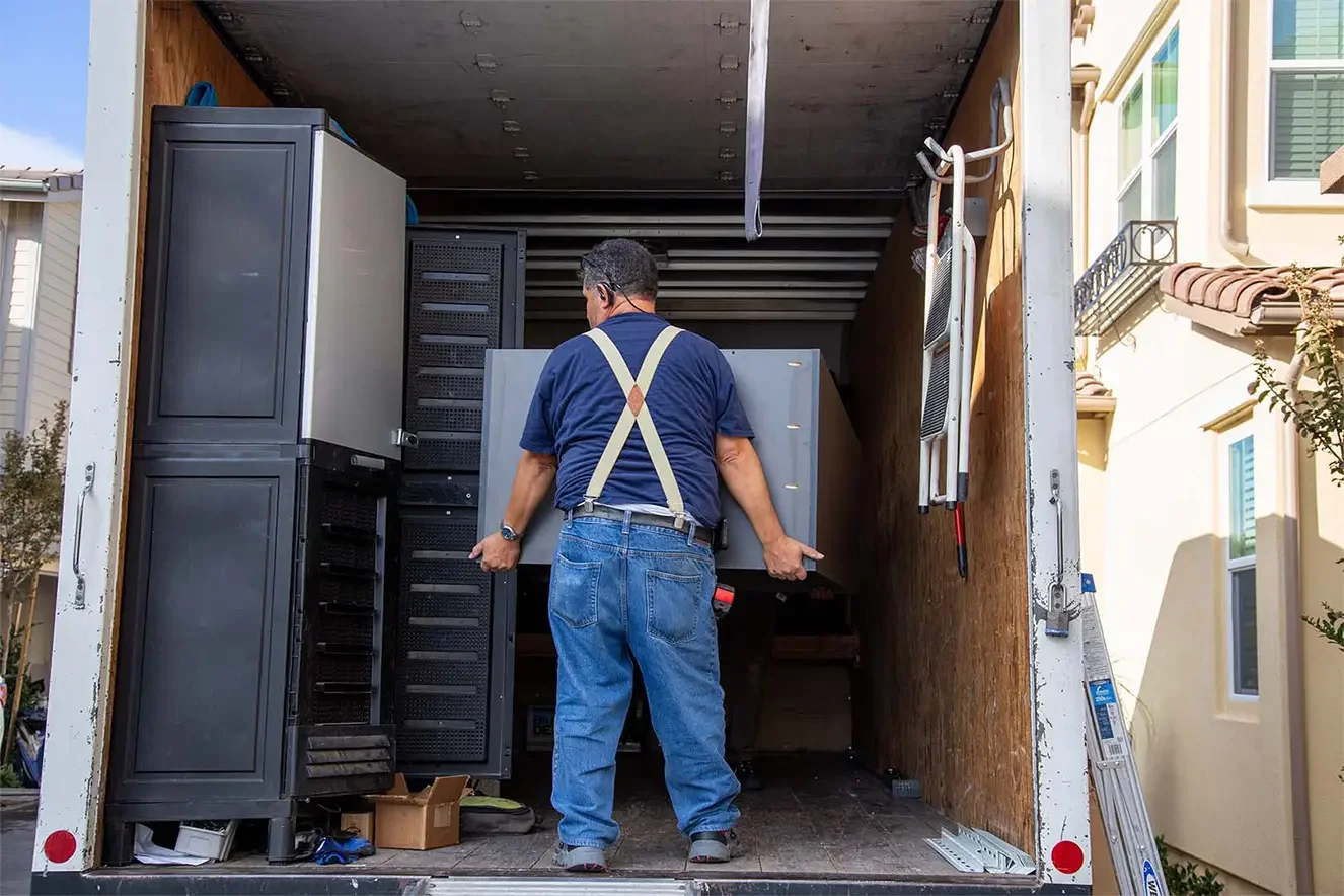 A man is moving a large light gray appliance into a moving truck. The truck's interior walls are wooden, and there are other appliances and boxes inside. The man is wearing blue jeans, a navy shirt, and suspenders.