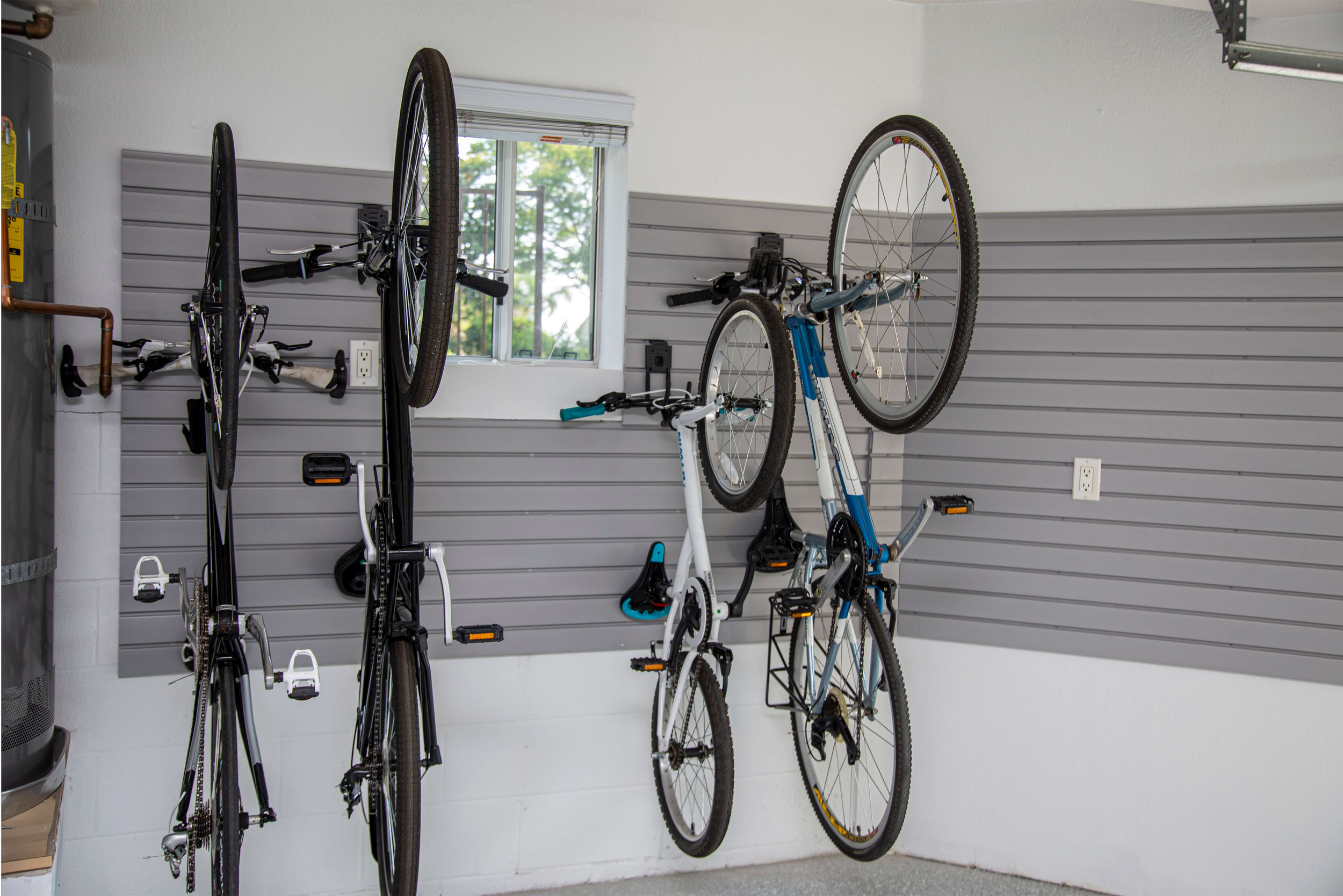 Indoor garage with gray slat wall mounted bike racks holding multiple bicycles of various sizes and styles, including mountain and hybrid bikes, in front of a small window with trees visible outside.
