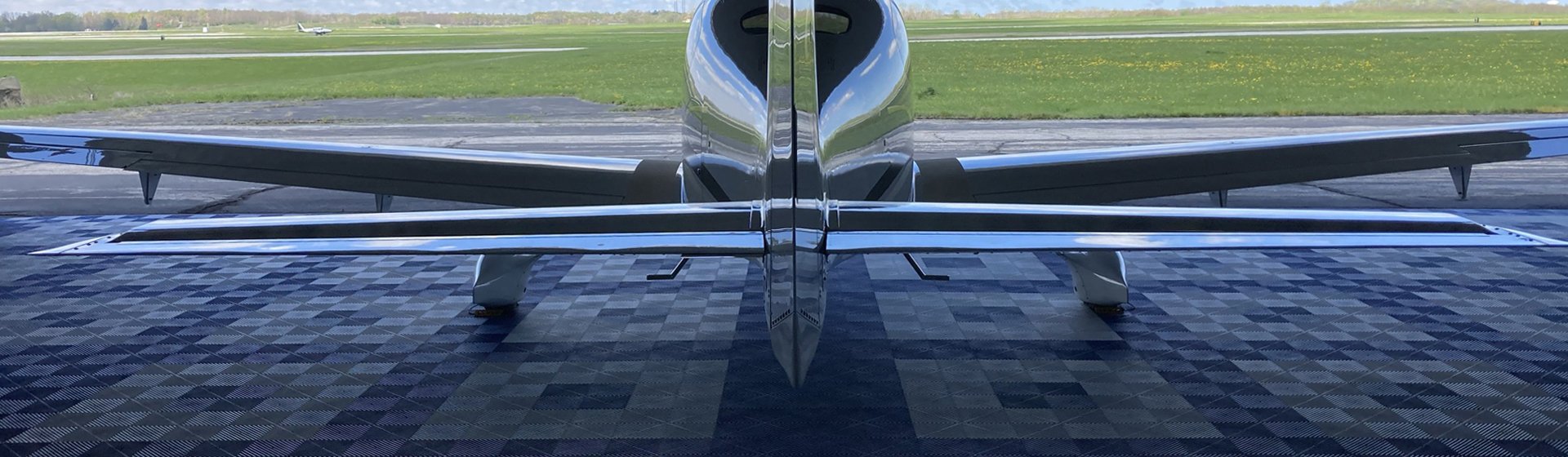 Front view of a small, sleek airplane parked at an airport gate with the runway and green fields in the background.