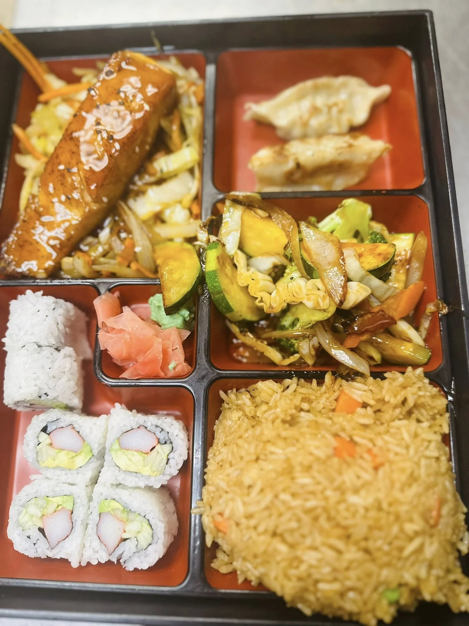 A bento box with assorted Japanese foods including sushi rolls, grilled vegetables, rice, fried rice, gyoza dumplings, and a stick of tempura.