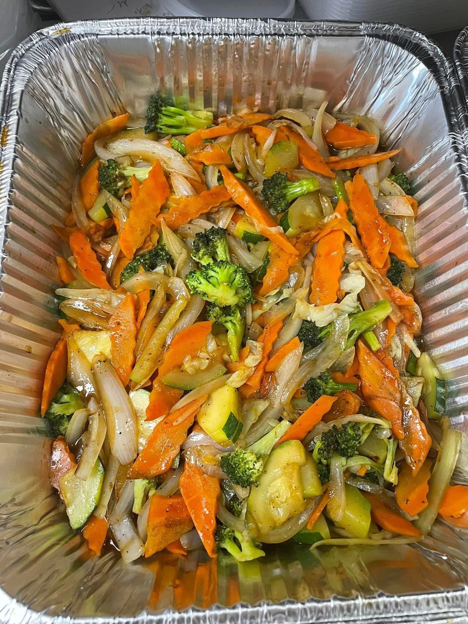 Mixed cooked vegetables including carrots, broccoli, onions, zucchini, and mushrooms in a foil container.