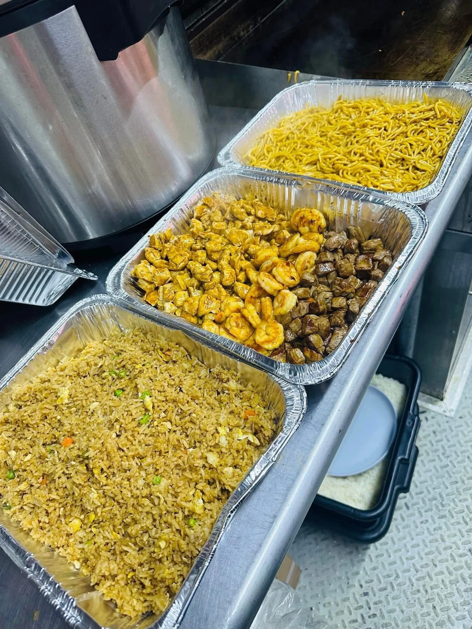 Aluminum trays filled with cooked Asian-style fried rice, stir-fried noodles, shrimp, and diced meat, placed on a counter in a food service setting.