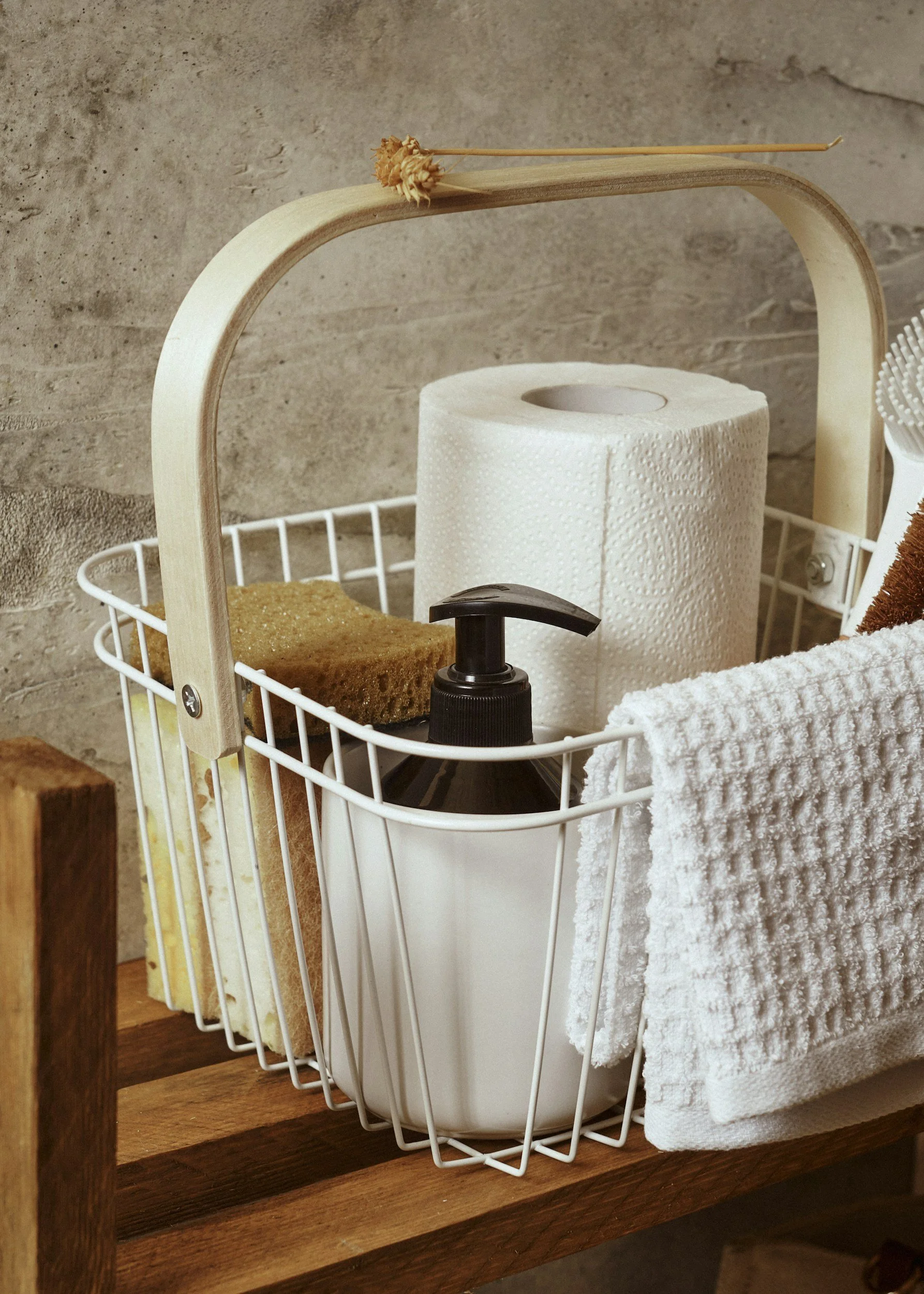 A white wire basket on a wooden surface holding a roll of paper towels, a bottle of soap or sanitizer, a sponge, and a white towel. There is a wooden handle on the basket and a small piece of dried plant or decor on top of the handle.