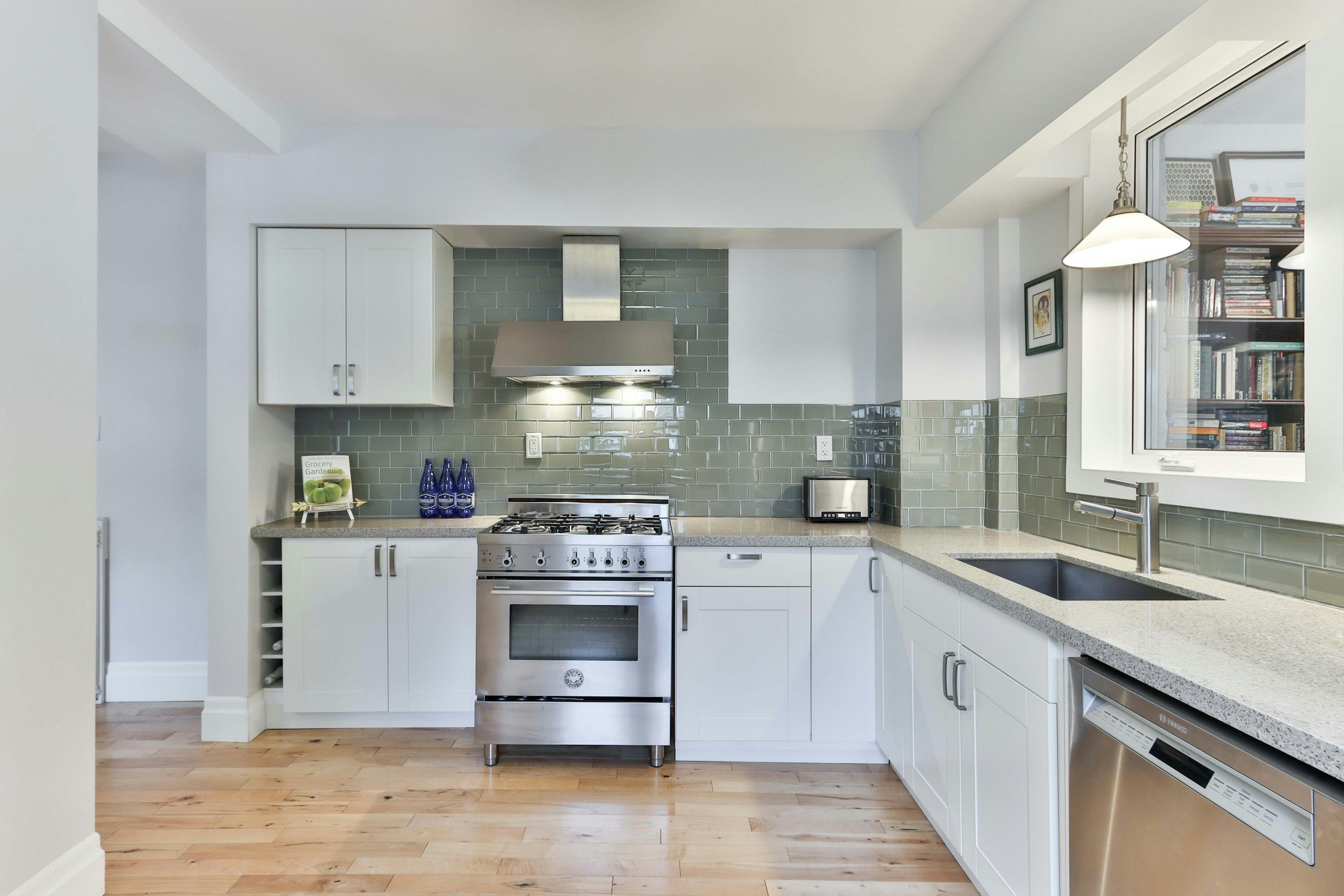 Modern kitchen with white cabinets, stainless steel stove, green subway tile backsplash, and a window with bookshelves outside.