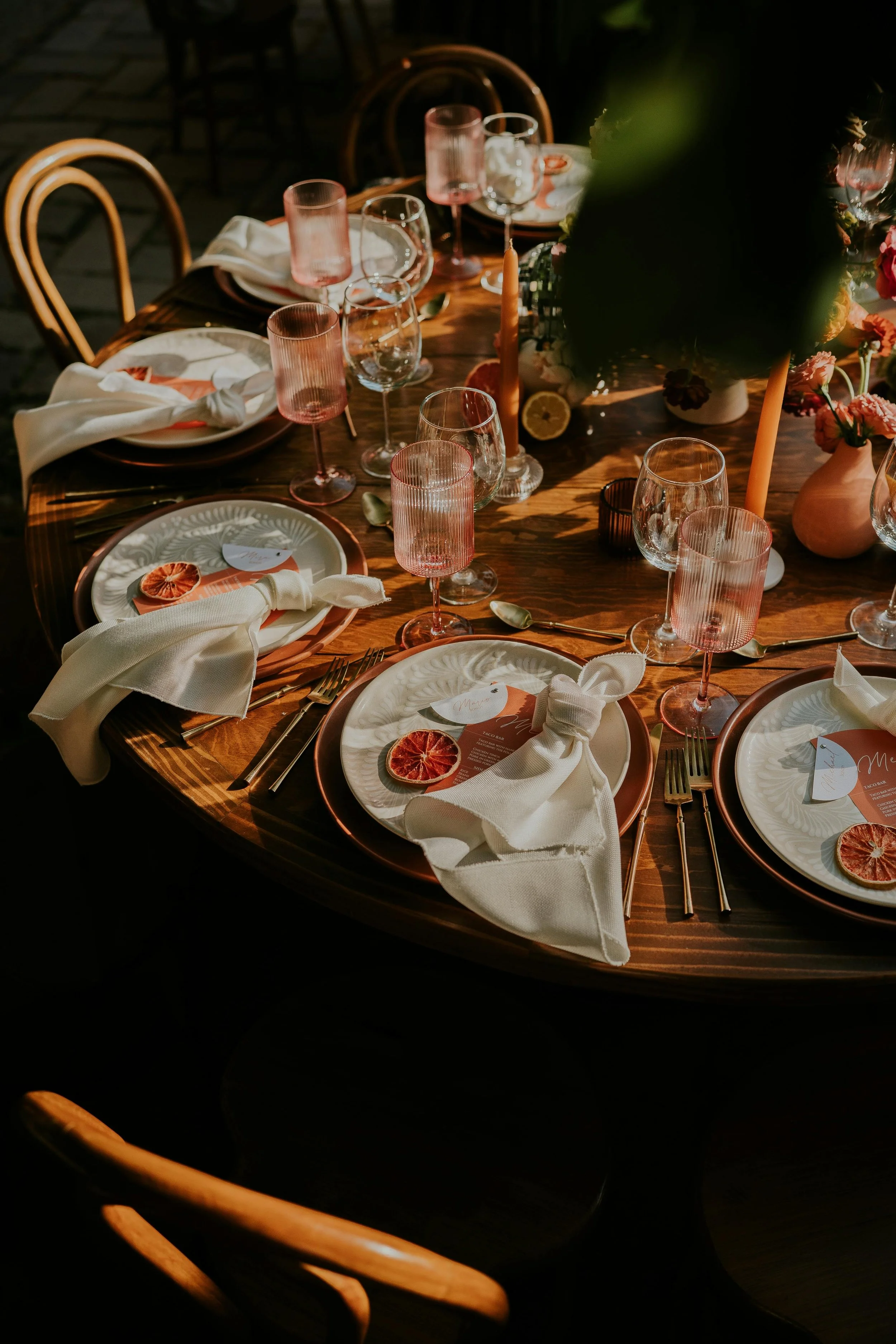 A formal dining table set for a meal with white plates, cream napkins tied with a ribbon, candles, wine glasses, and floral centerpieces, illuminated by warm lighting.