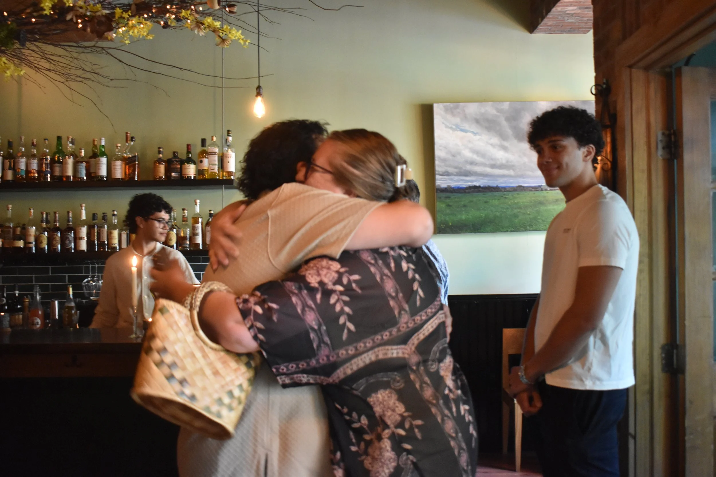 A group of people hugging inside a bar restaurant with bottles of alcohol on shelves in the background. A man behind the bar is preparing drinks, and a young man is standing nearby, smiling.