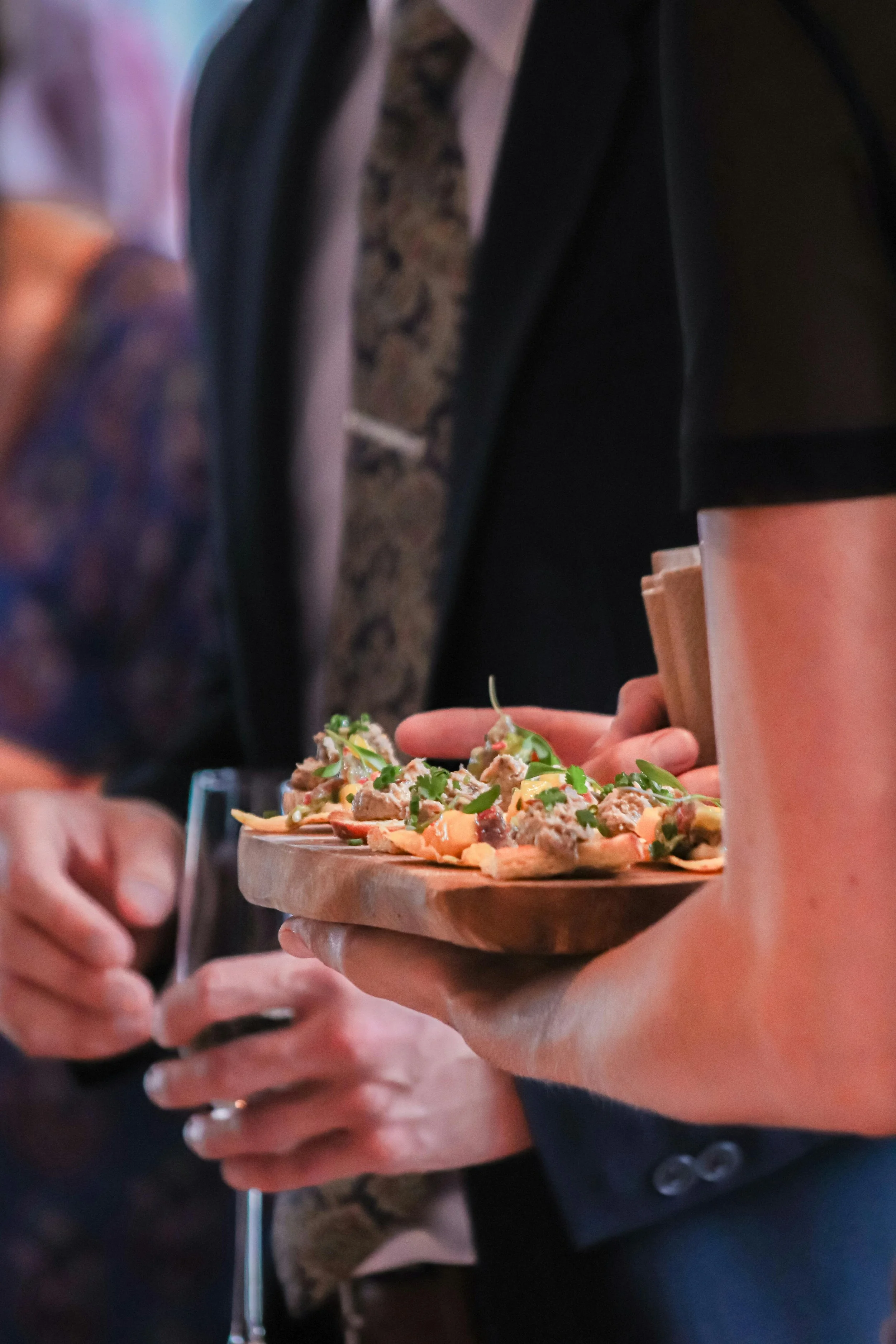 Person holding a wooden serving board with a pizza topped with green herbs and various ingredients, at a social gathering or restaurant.