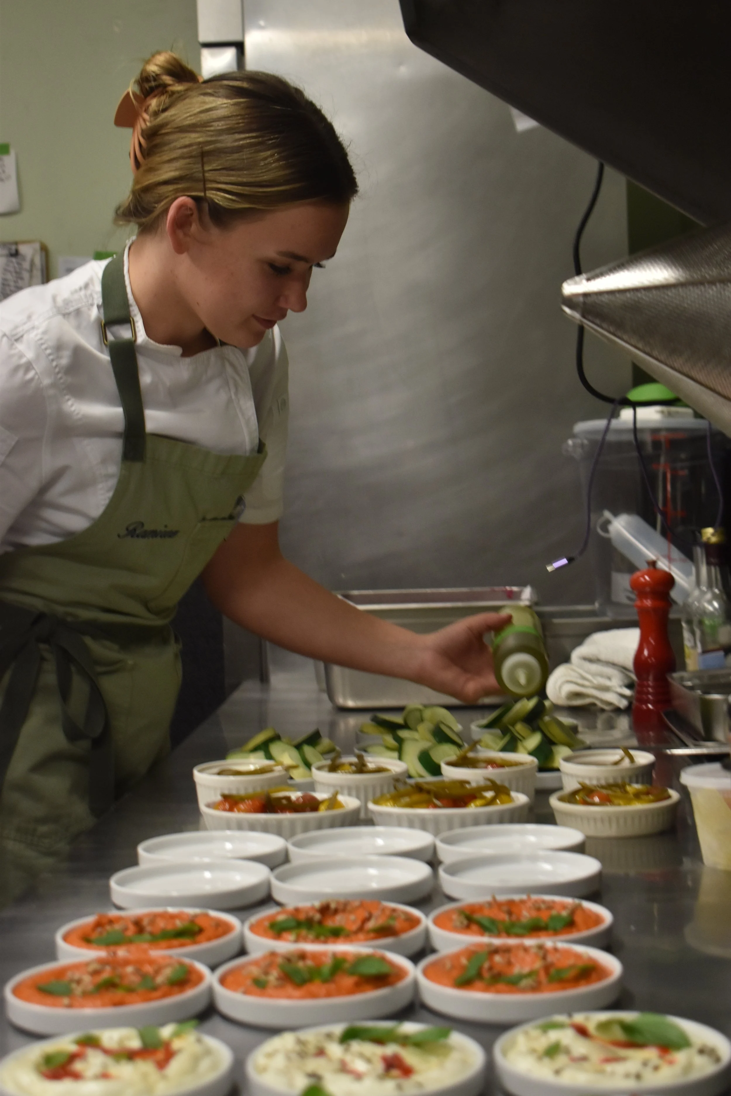 A woman in a chef uniform and apron preparing food in a commercial kitchen, with bowls of prepared food and ingredients on a stainless steel countertop.