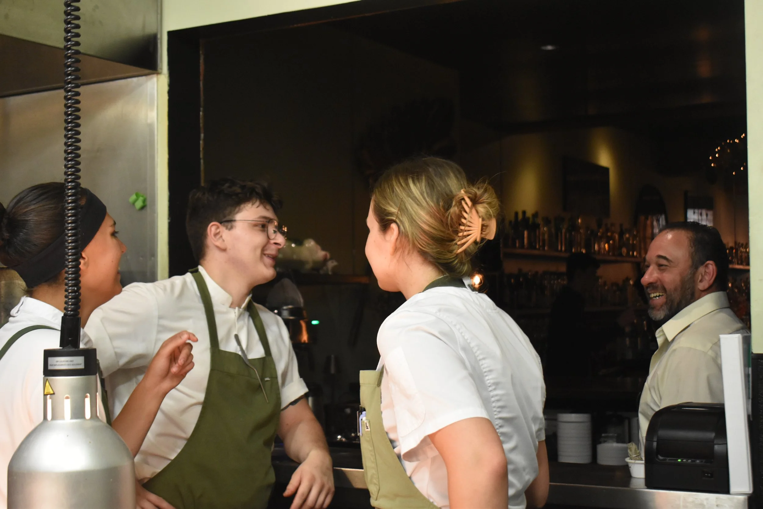 Group of four people wearing white shirts and green aprons, standing in a kitchen, engaging in conversation. Three women and one man, with one woman having her hair tied up with a hair clip and another wearing a black headband. The background includes kitchen shelves with bottles and glasses and a darkened room.