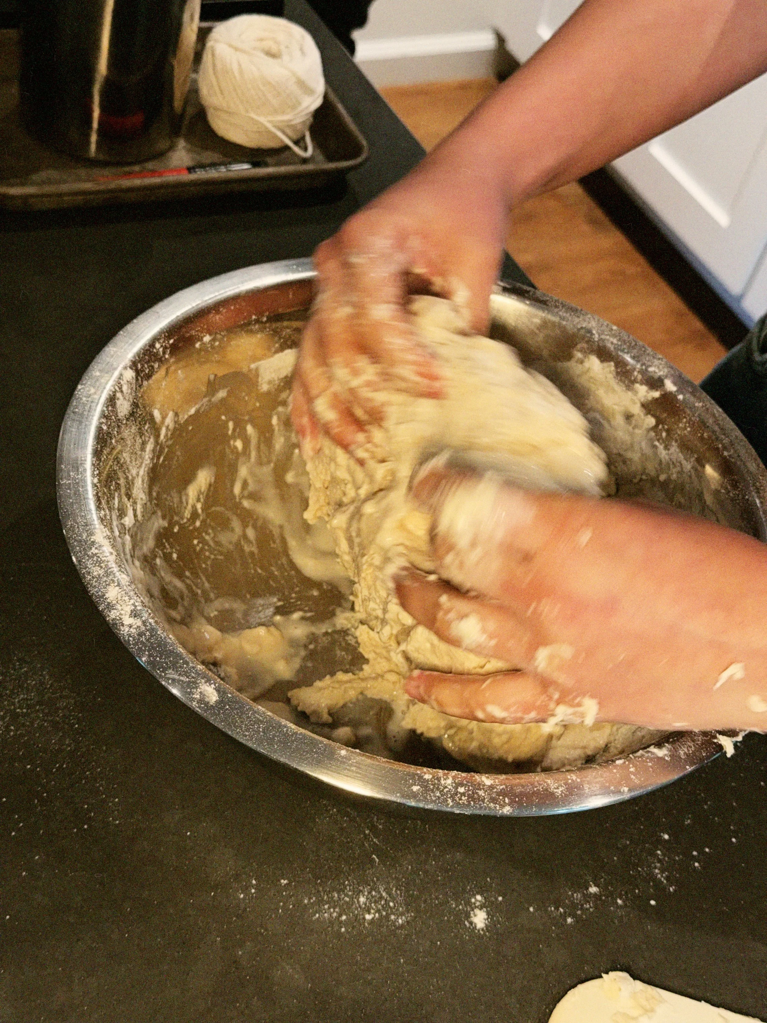 Hands kneading dough in a metal mixing bowl on a black countertop, with some flour sprinkled on the surface.