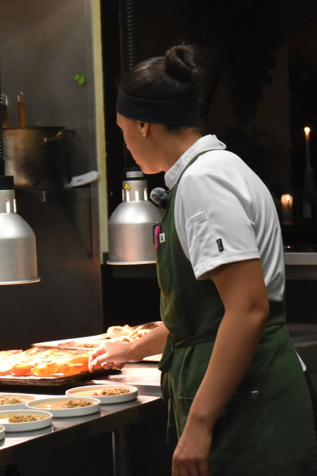 A woman wearing a black headband and white shirt with a green apron is preparing food behind a counter with plates of dishes and kitchen equipment.