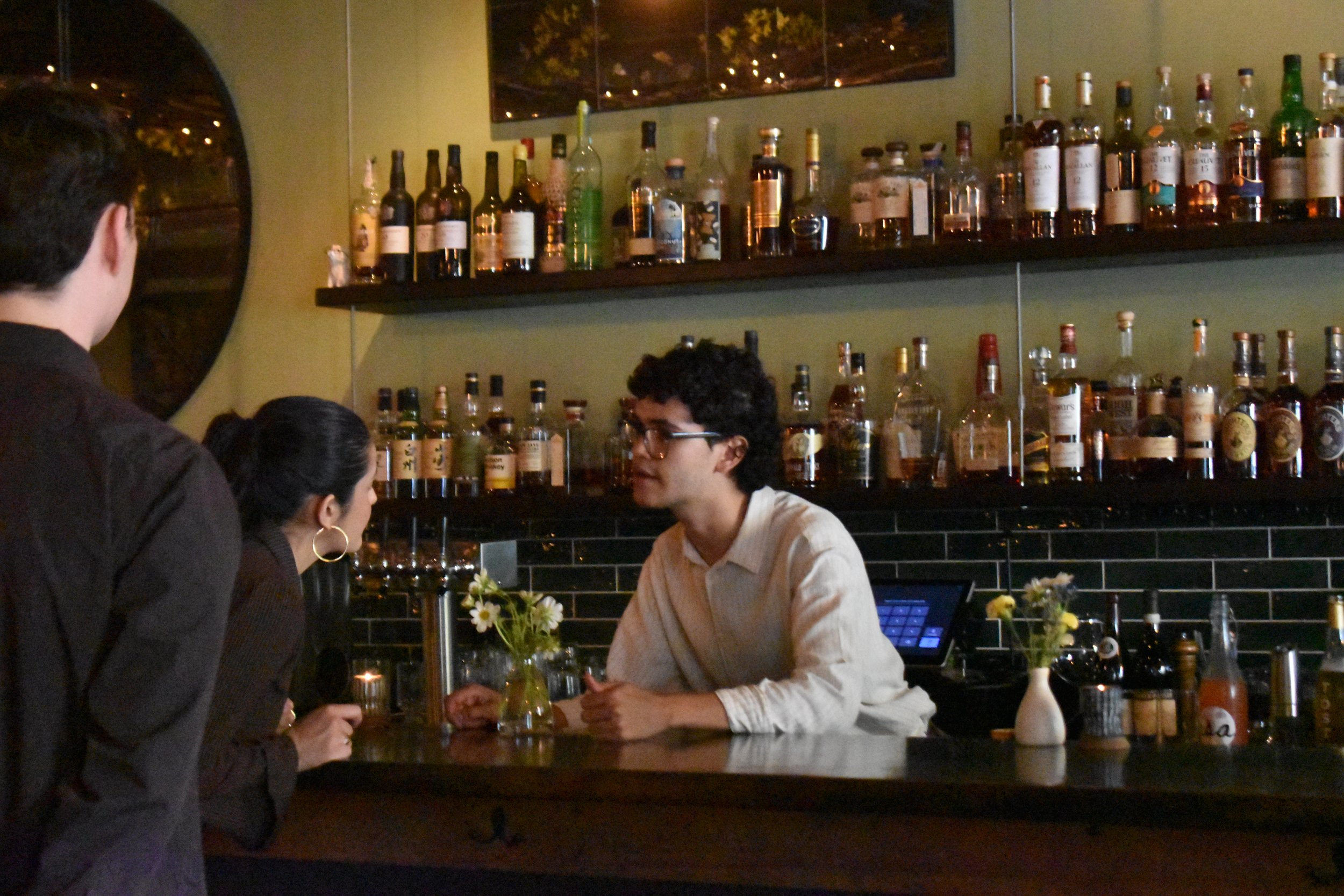 A bartender interacts with three customers at a bar, surrounded by shelves of liquor bottles and floral decorations.